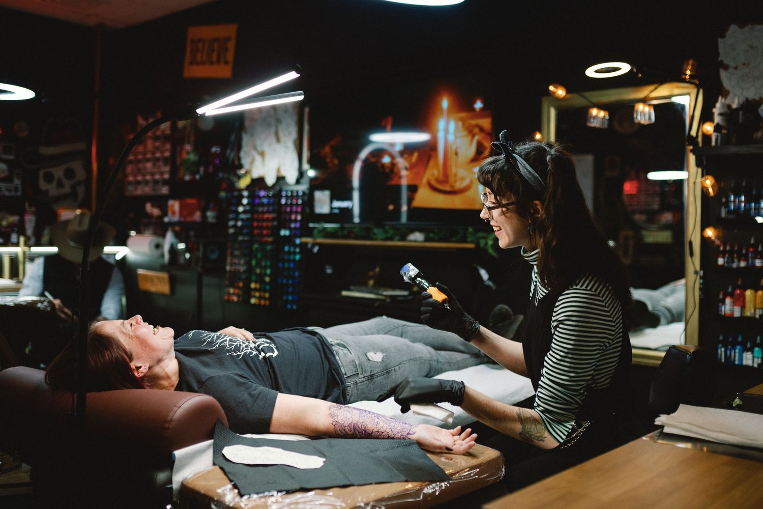 Tattoo artist works on client's arm in dimly lit studio with professional equipment and supplies.