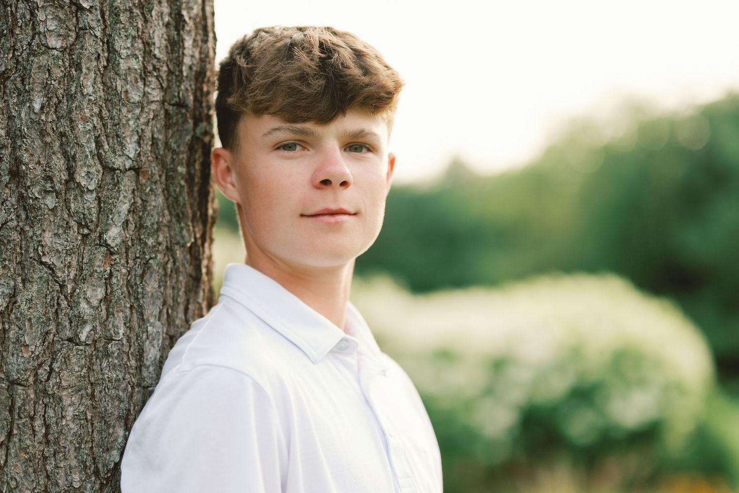 Teen boy in white shirt leaning against tree trunk outdoors in soft natural light.