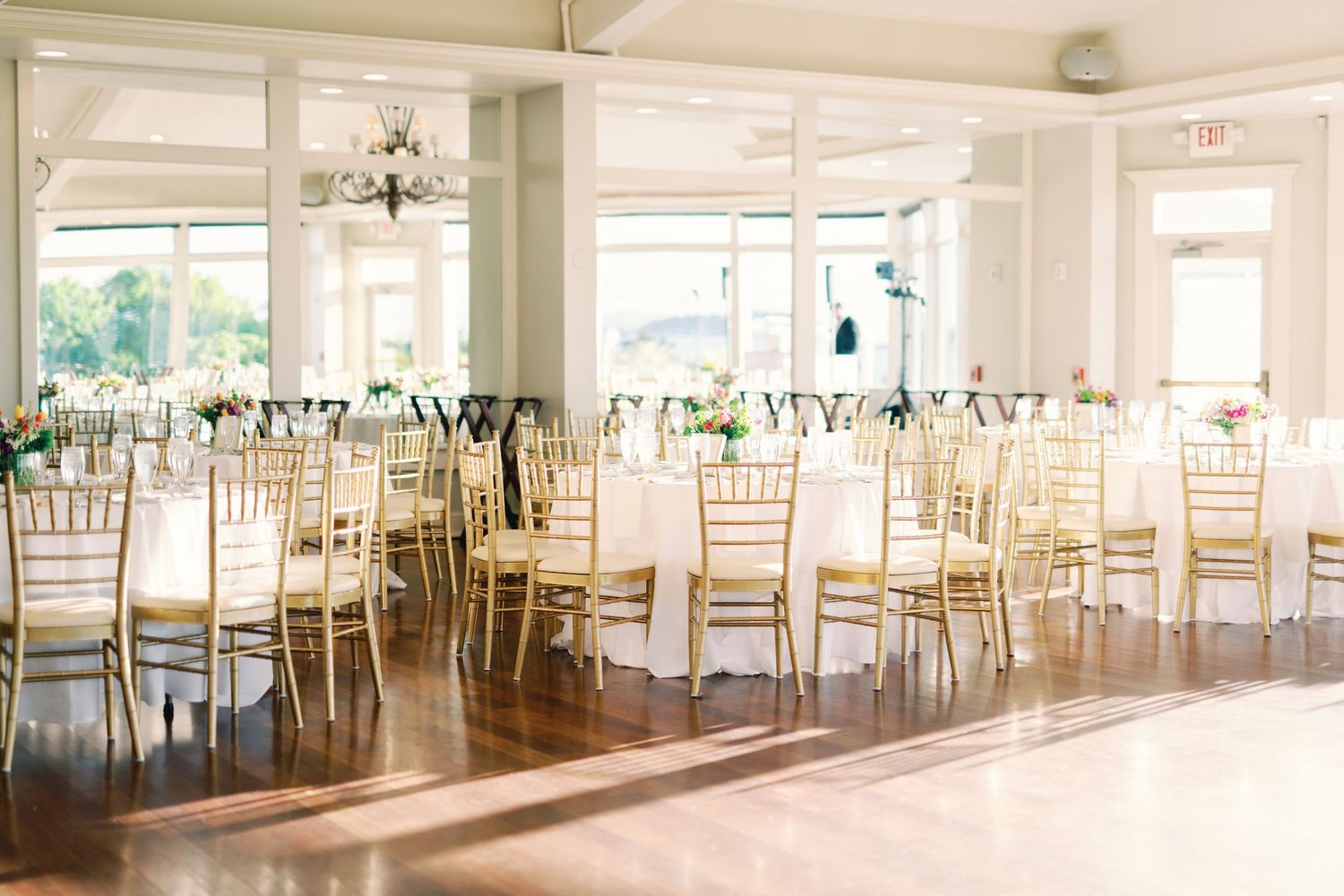 Elegant wedding reception venue with gold chiavari chairs and white linens in a bright room with hardwood floors.