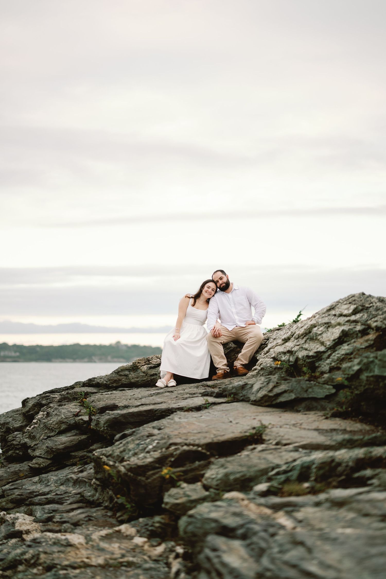 A couple in elegant attire sits together on coastal rocks during an overcast evening by the water.