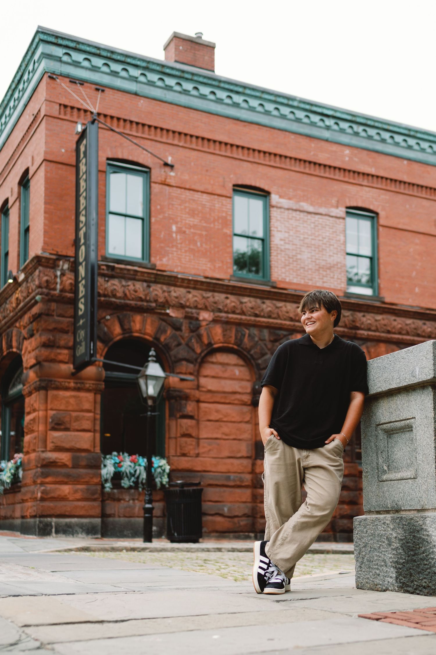 A person in black shirt and khaki pants leans against a stone pillar outside a historic red brick building with green trim.
