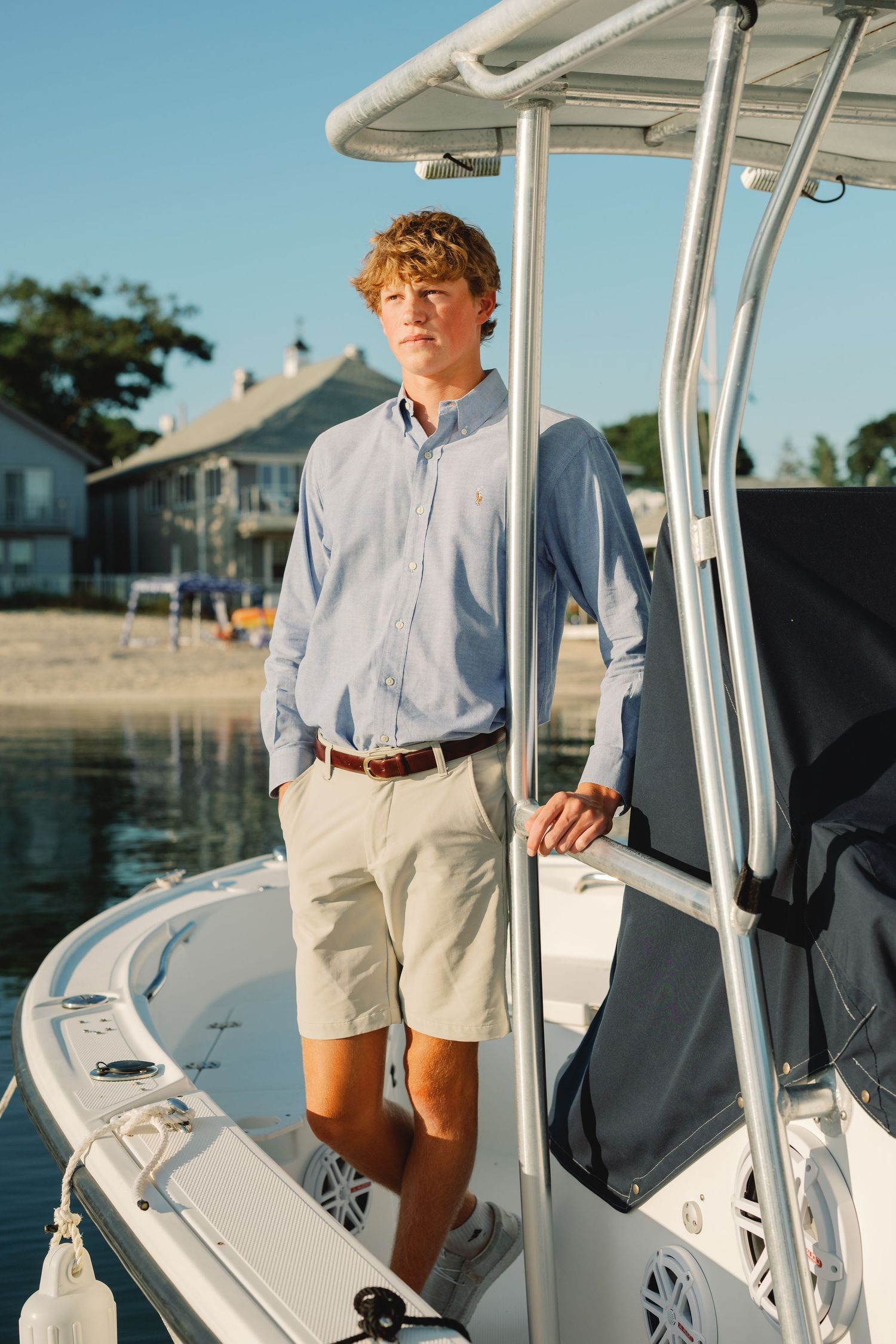 Young person in light blue shirt and khaki shorts standing on a white boat at sunset by the waterfront.