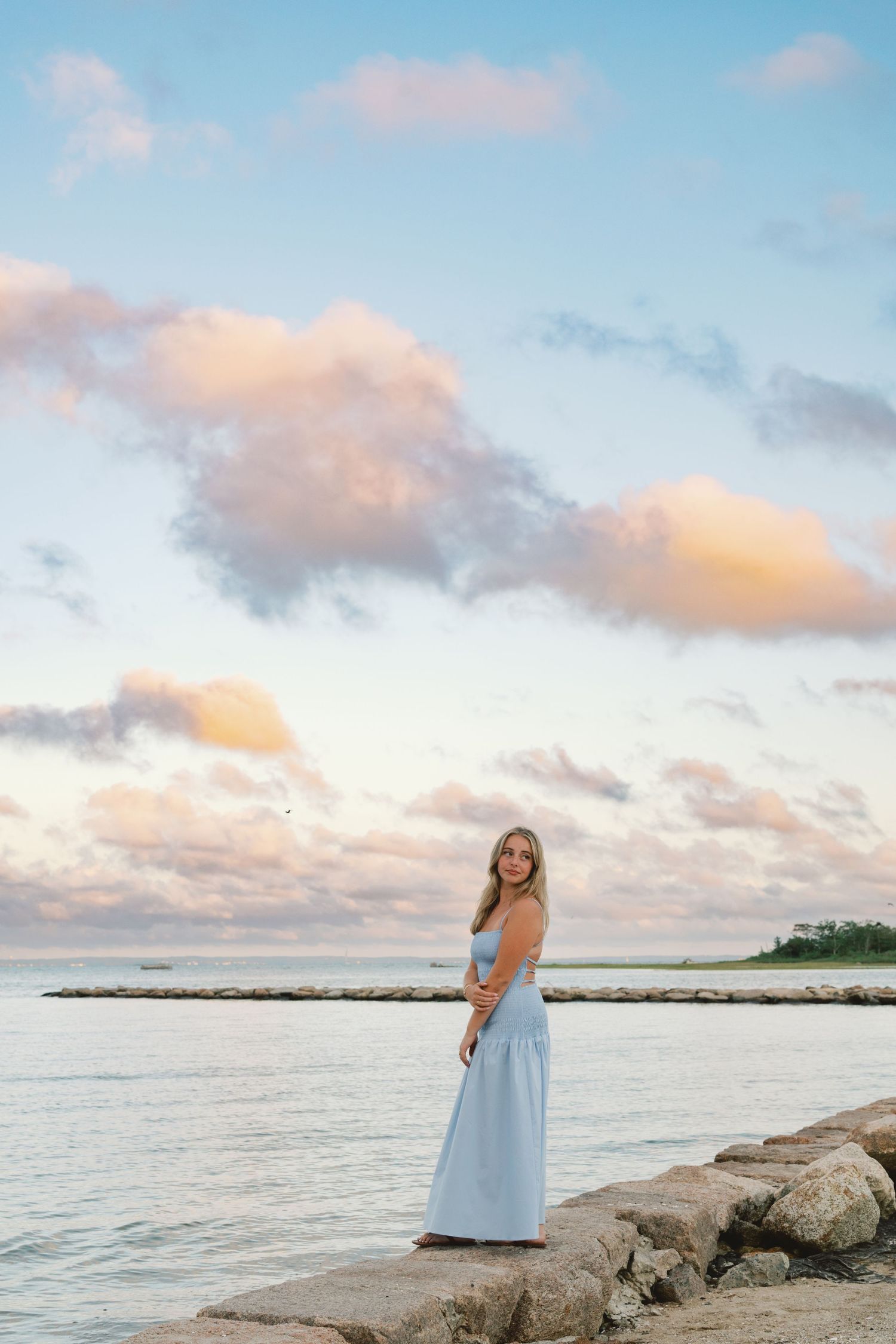 A person in a light blue dress stands on a rocky shoreline at sunset with pink clouds and calm waters in the background.