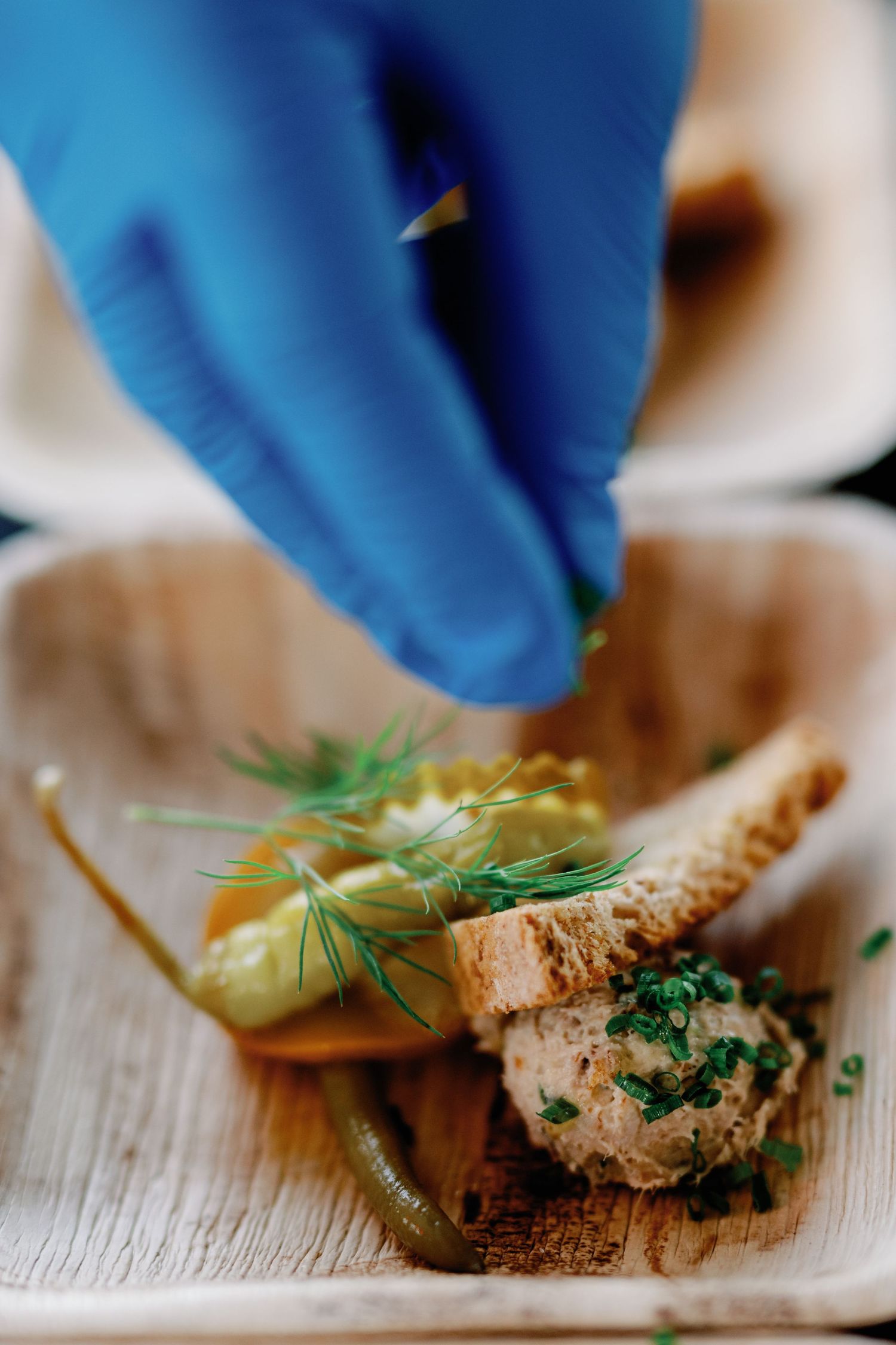 A gloved hand garnishes food with fresh dill and herbs on a wooden cutting board.