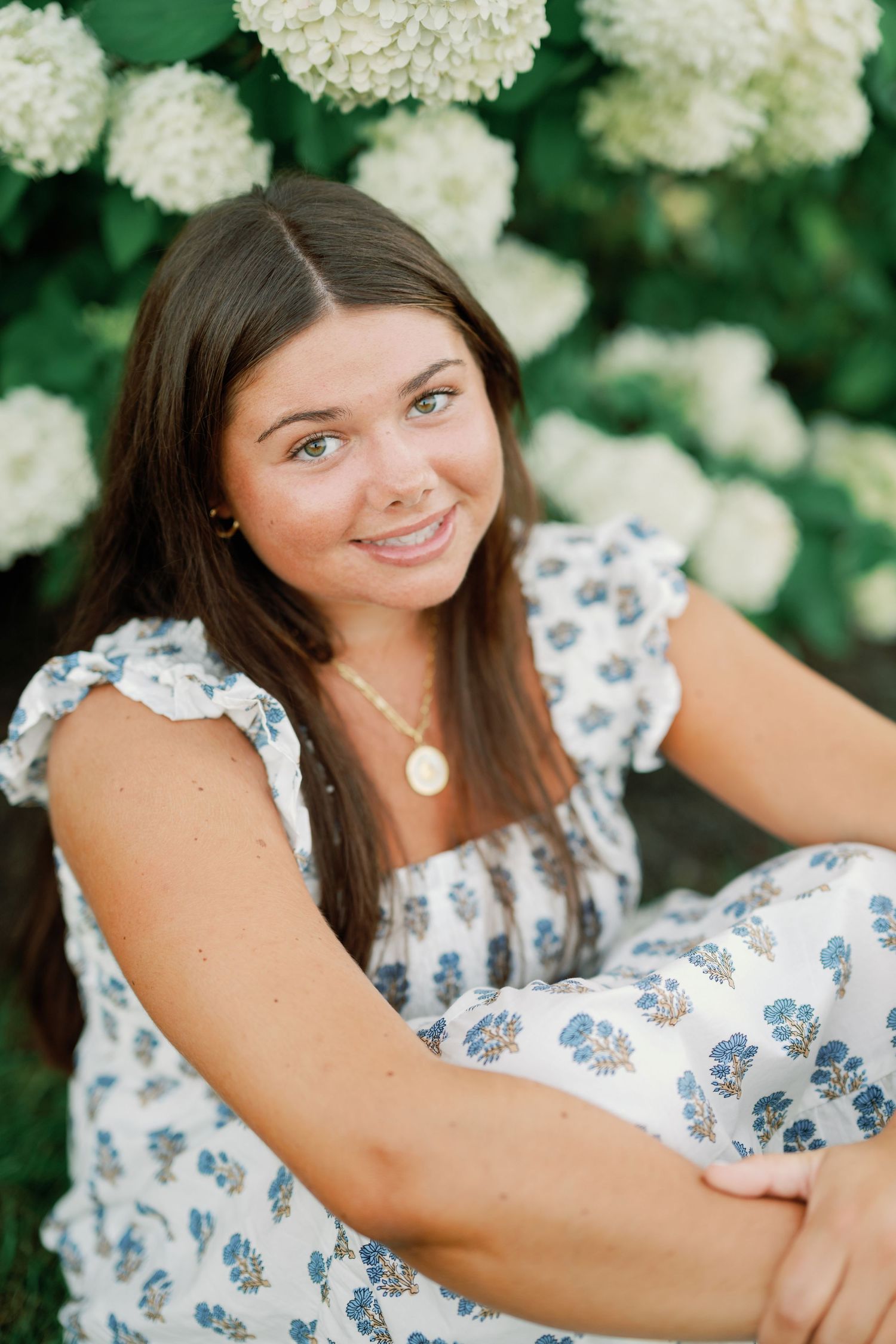 Young woman in floral dress sitting outdoors among white hydrangea blooms, smiling warmly at camera.