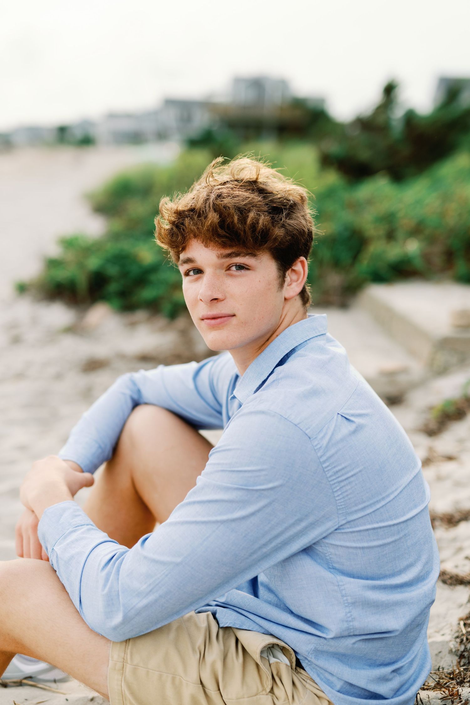A person in a light blue button-down shirt and khaki shorts sits casually on a rocky beach.