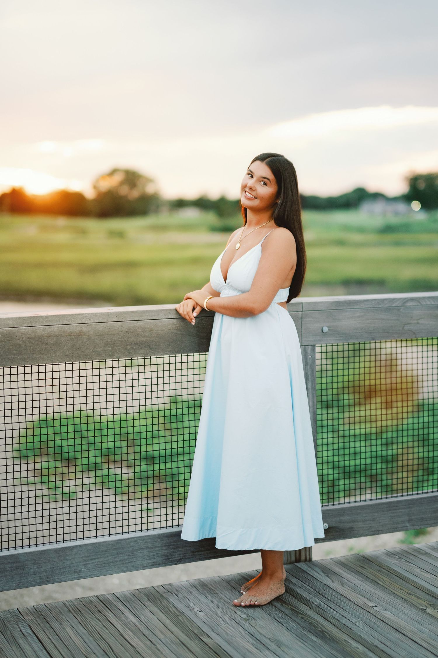 Young woman in white midi dress standing barefoot on wooden deck at sunset, smiling softly at camera.