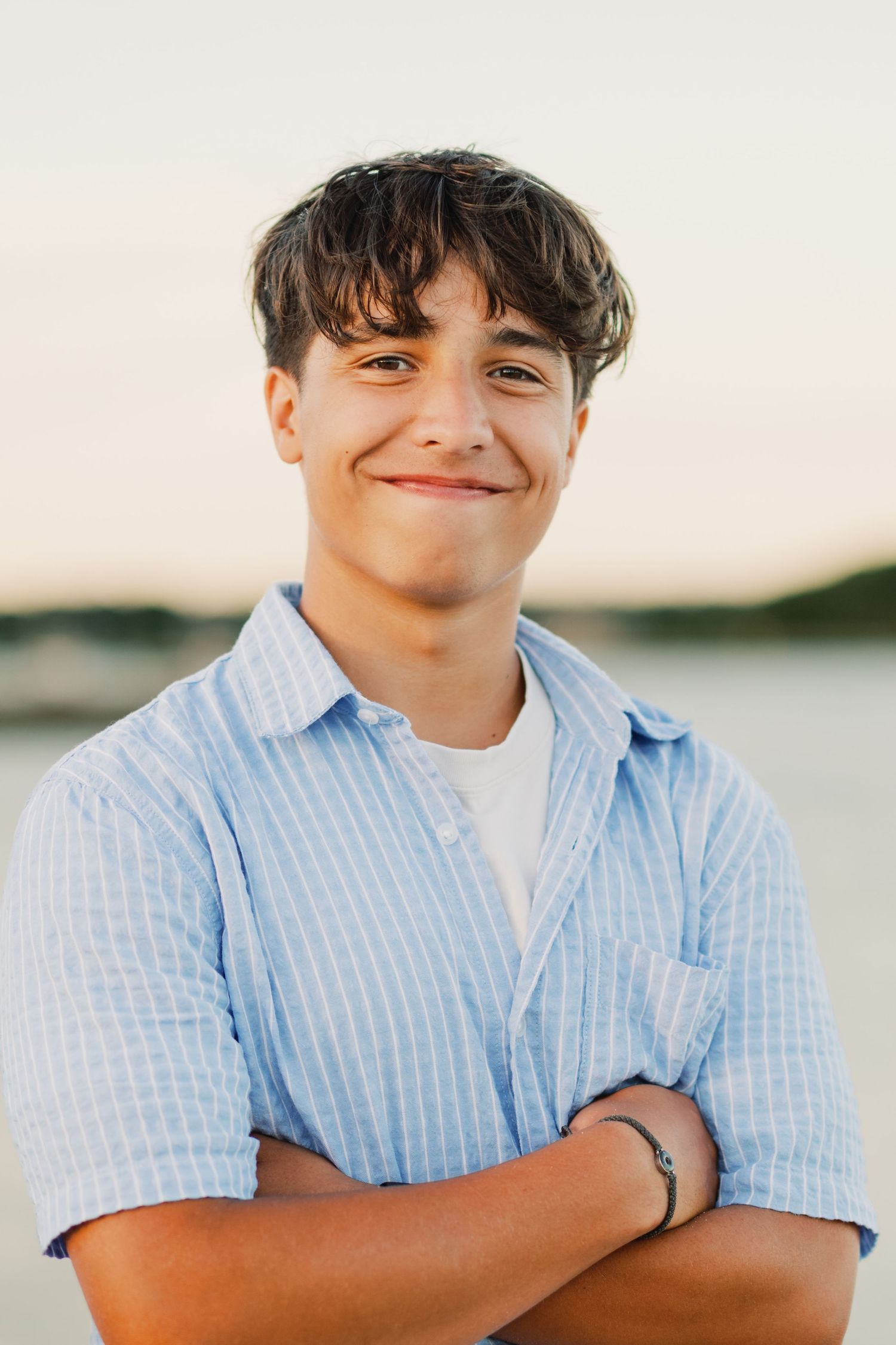 A smiling teenage boy with dark hair stands arms crossed wearing a light blue striped shirt near a waterfront at sunset.