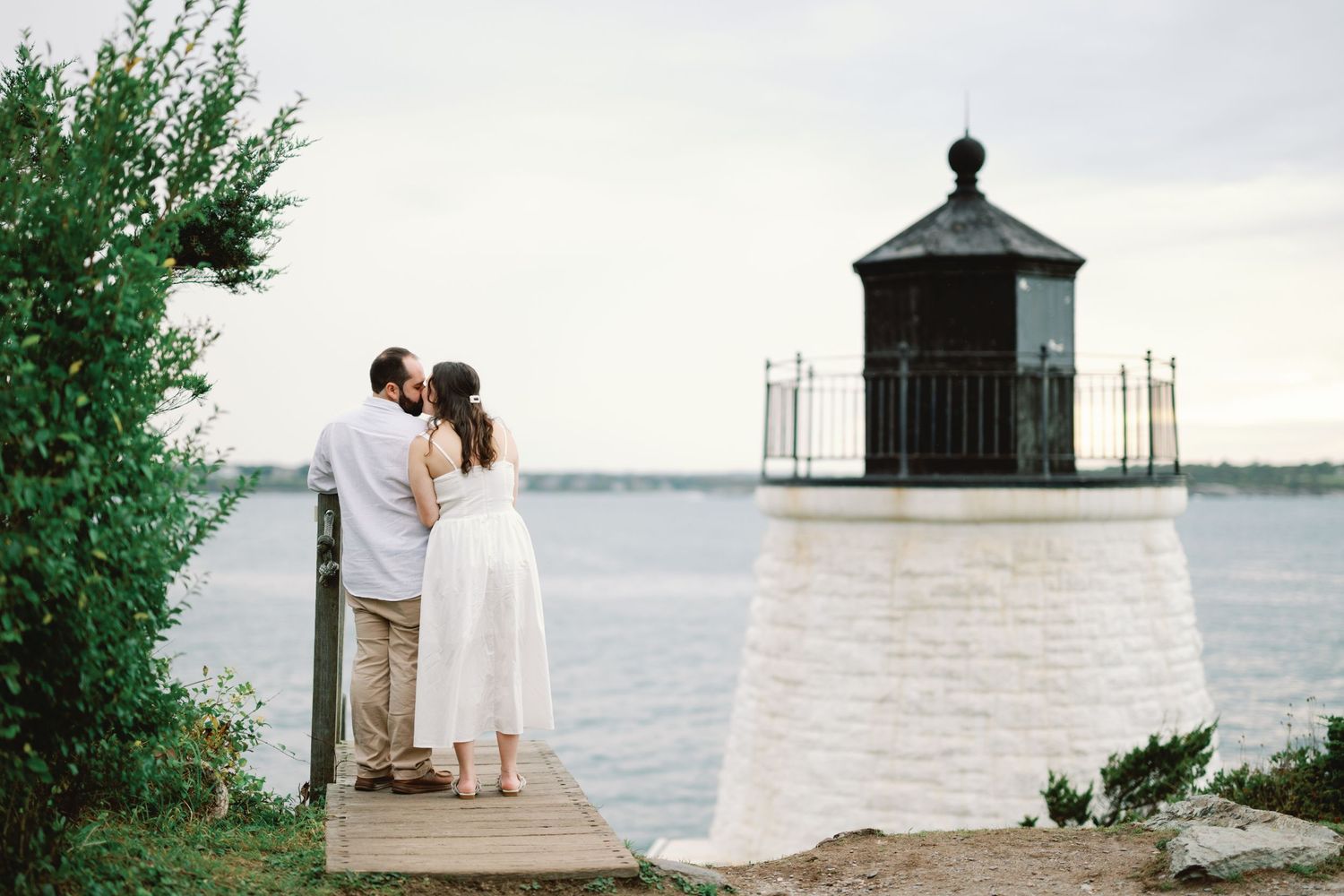 A romantic couple stands by a small white lighthouse overlooking a calm waterfront at dusk.