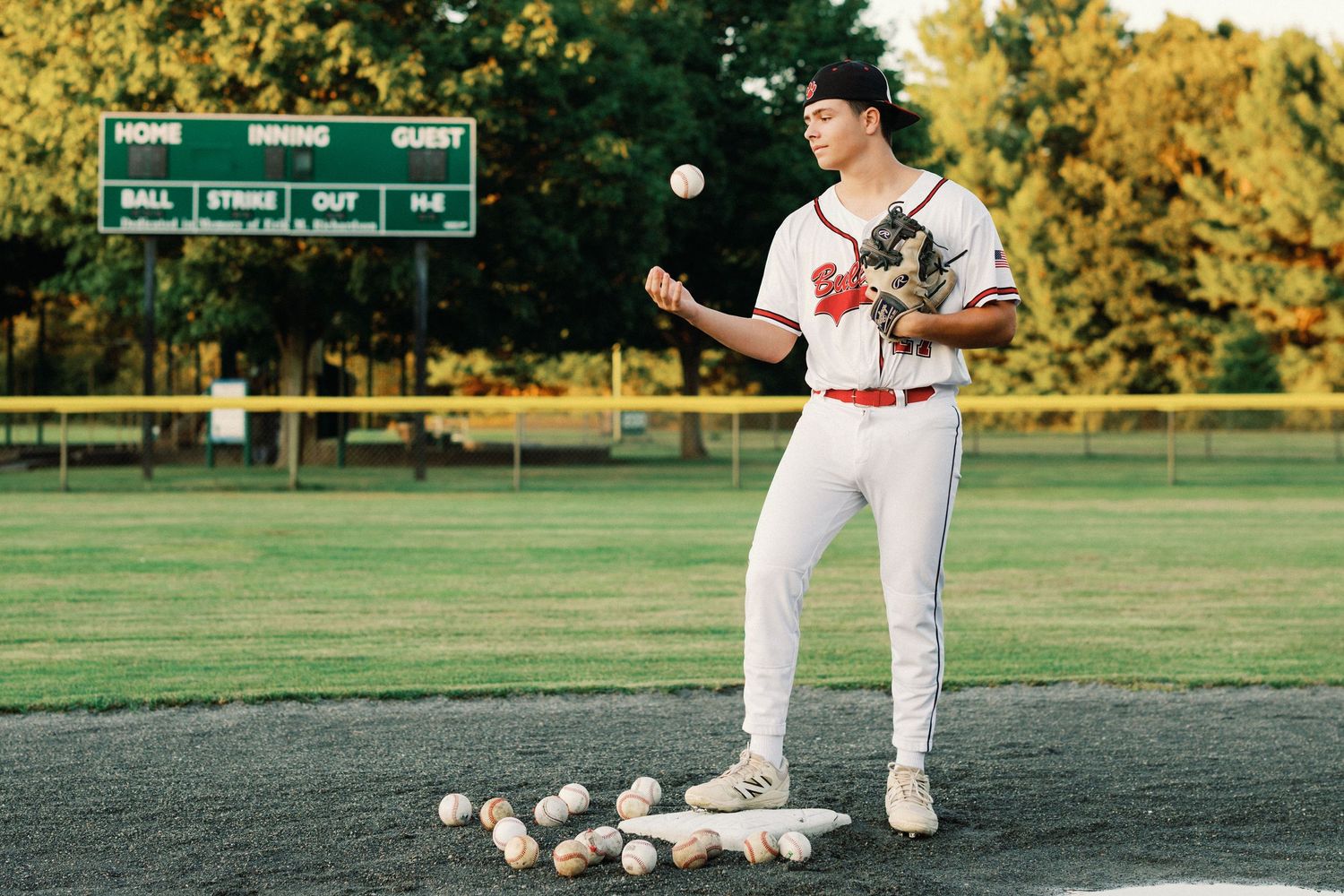 Baseball pitcher in white uniform tosses a ball on the mound surrounded by baseballs with a scoreboard in background.