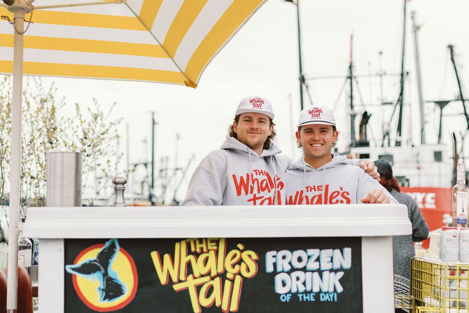 Two people stand behind a food cart with a yellow and white striped umbrella at a marina.