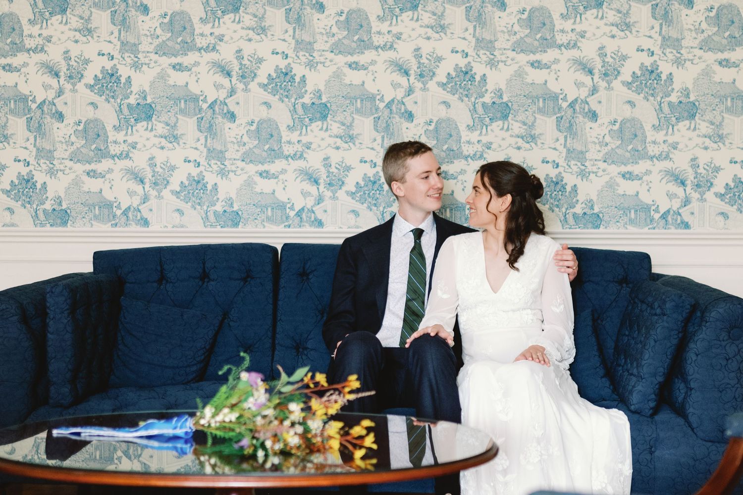 A couple in white attire sits on a blue velvet couch in front of a floral wallpapered wall.