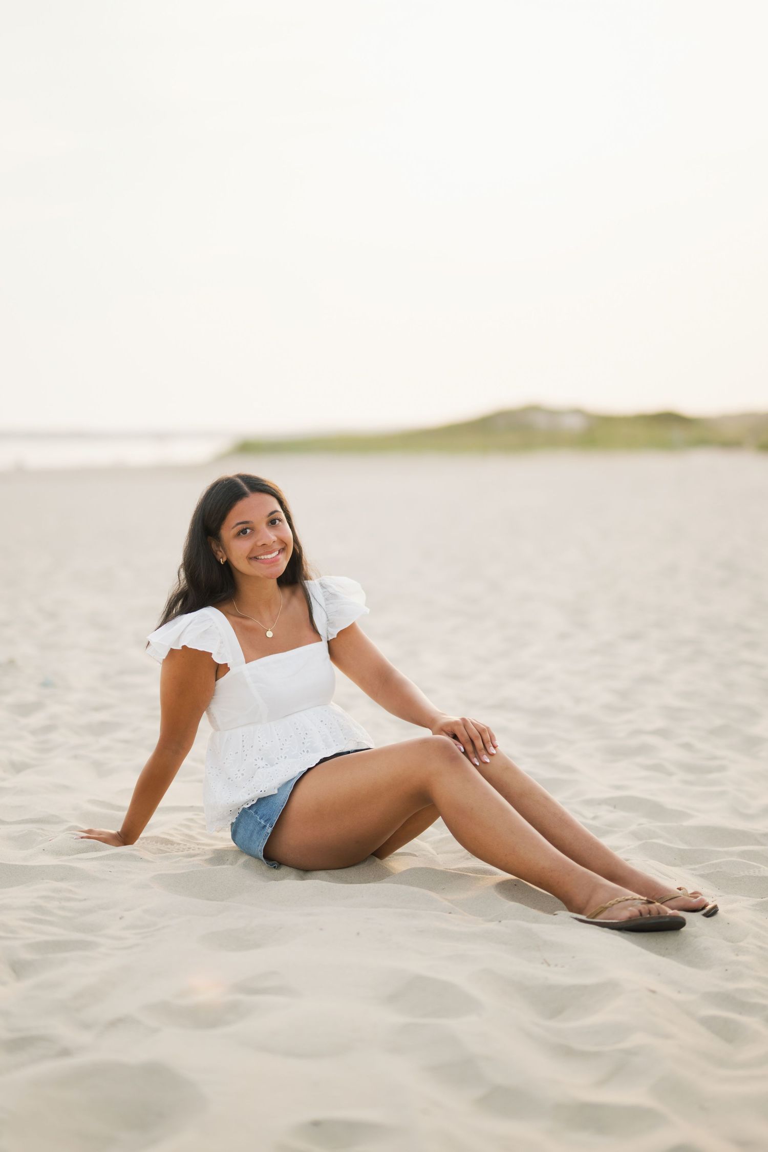 Young woman in white top and denim shorts smiling while sitting on sandy beach at golden hour.
