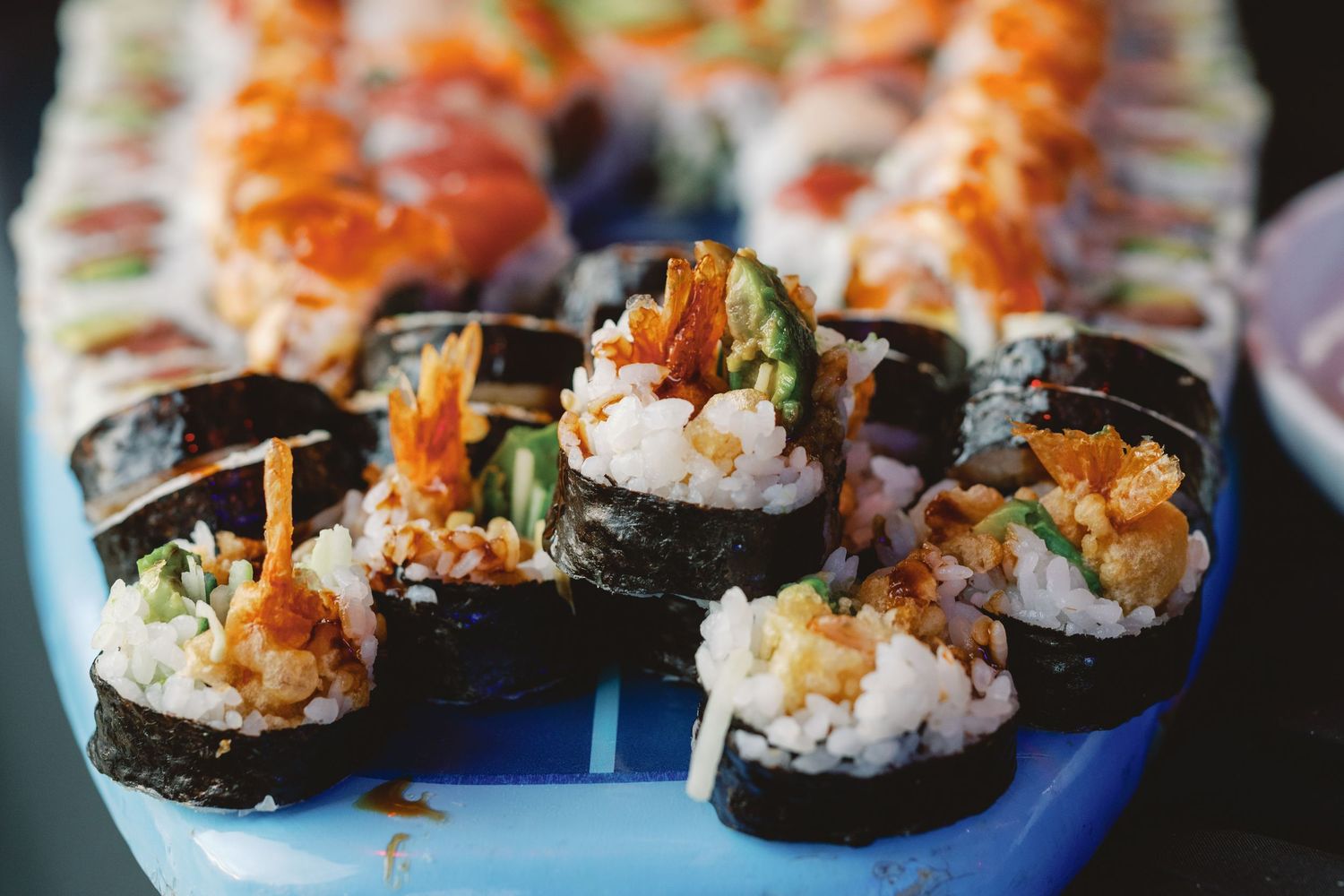 Freshly prepared sushi rolls with rice and fillings being arranged in a sequence on a blue surface.
