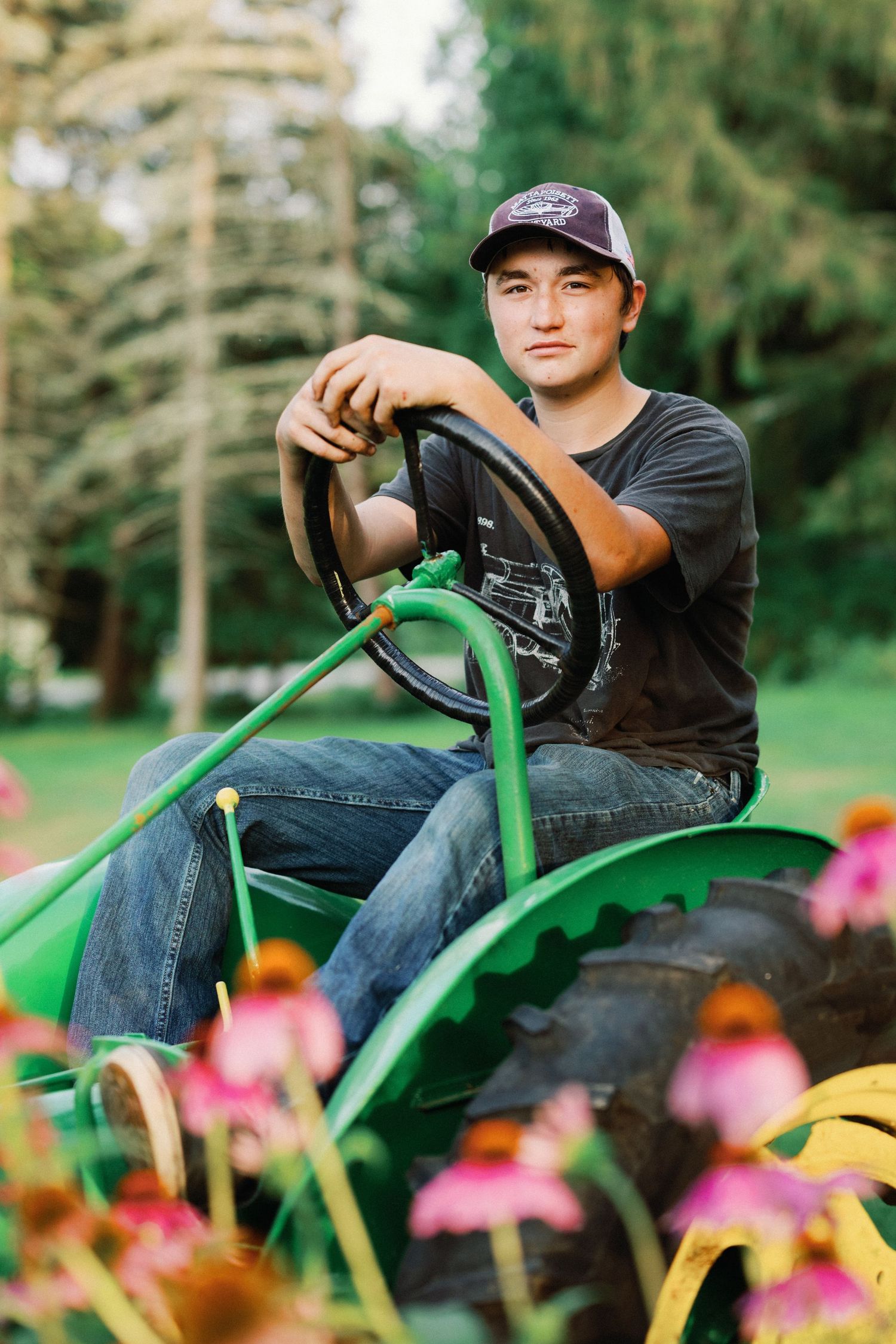 Young man in cap sitting on green John Deere tractor surrounded by pink flowers and trees.