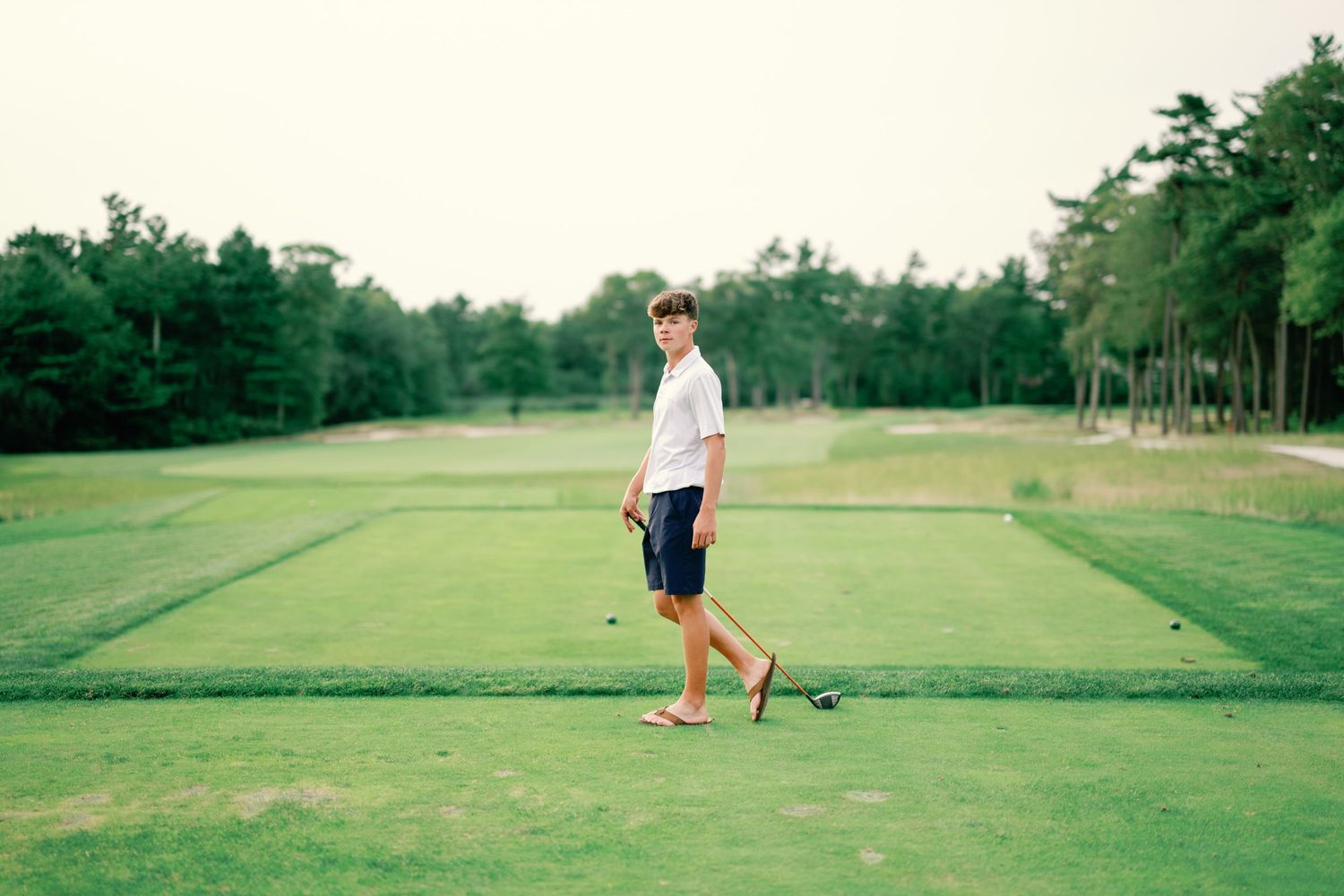 Young golfer standing on lush green golf course holding club, surrounded by trees on a clear day.