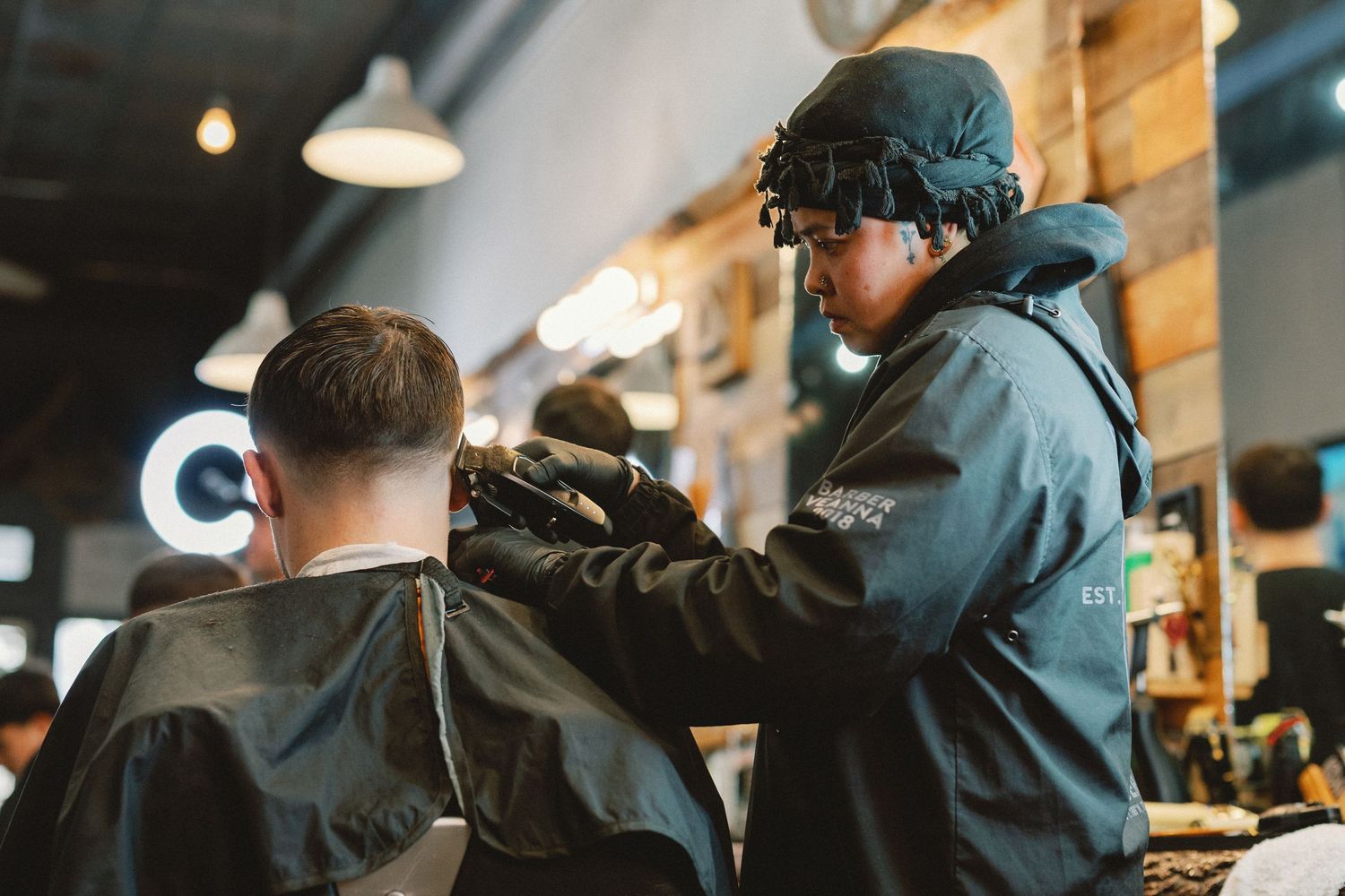 A barber uses professional clippers to trim a client's hair in a modern barbershop with warm lighting.
