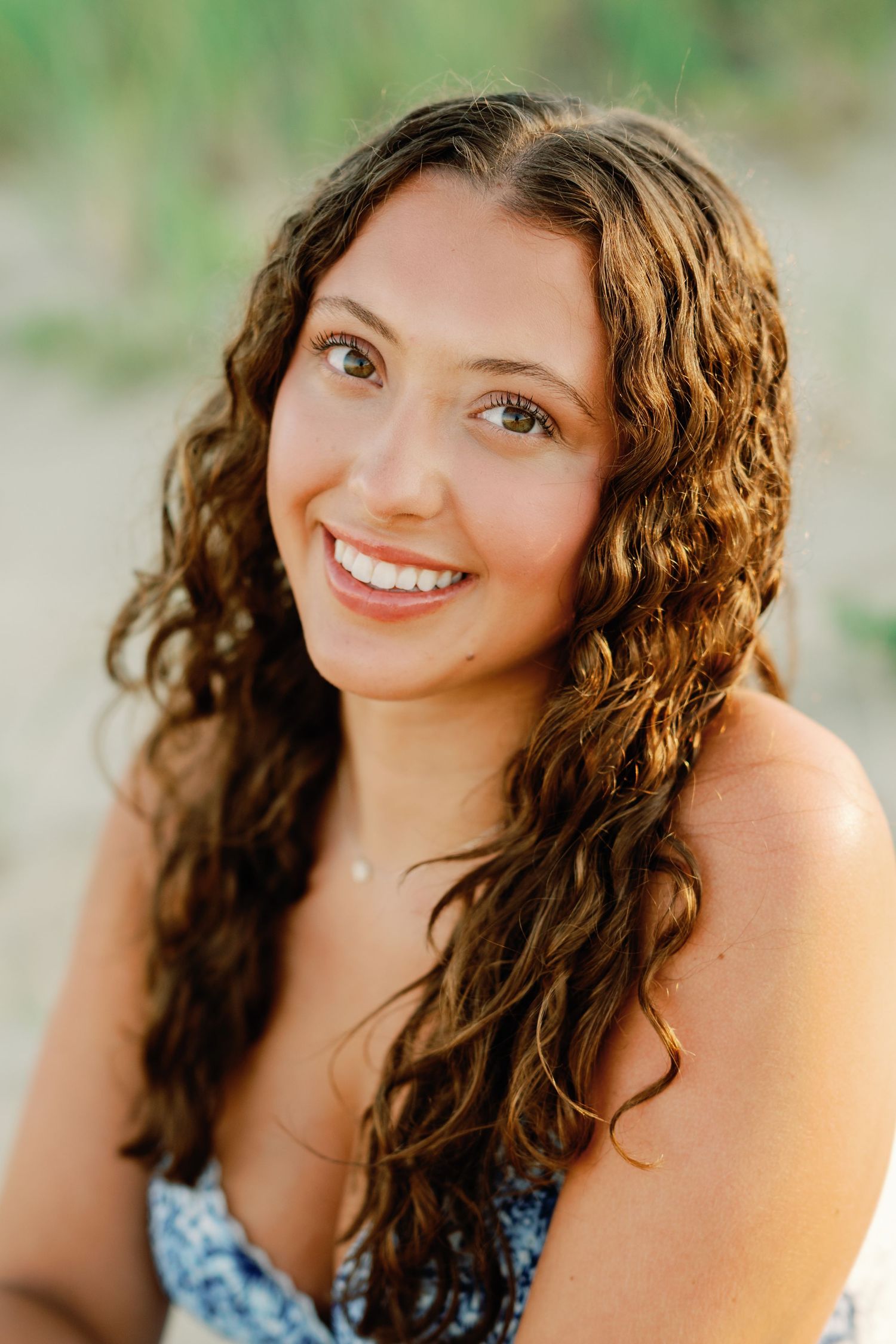 Young woman with curly brown hair smiling outdoors at the beach on a sunny day.