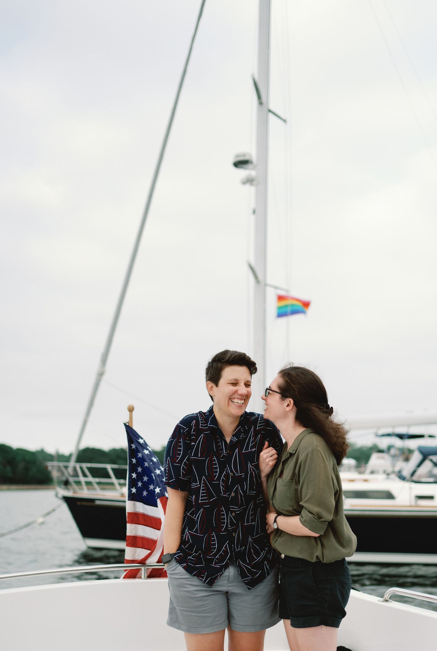 A couple shares a joyful moment on a sailboat with an American flag and boats visible in the marina background.