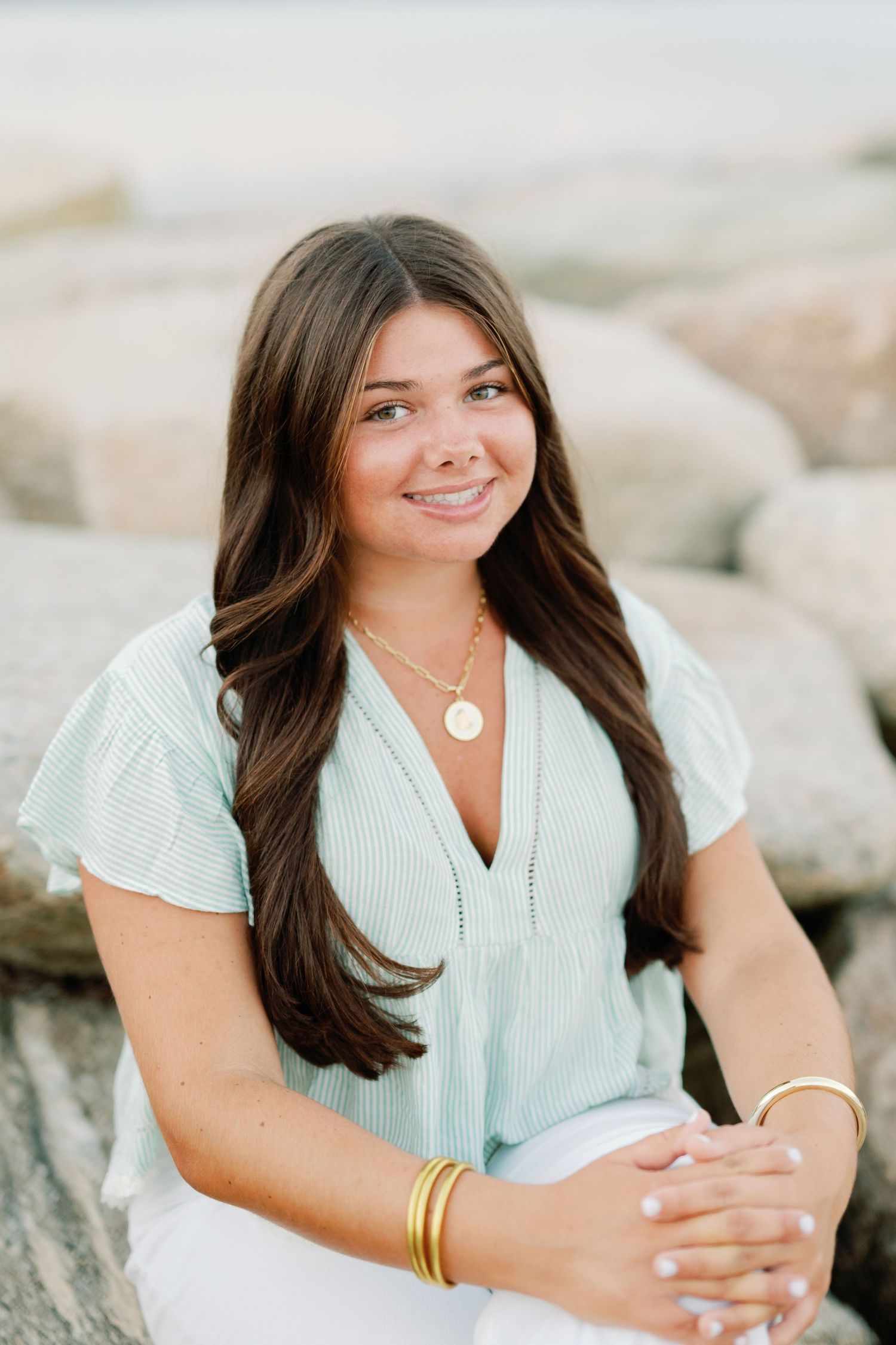 Young woman with long brown hair smiling outdoors, wearing a mint top, gold necklace, and bracelets, seated on rocks.