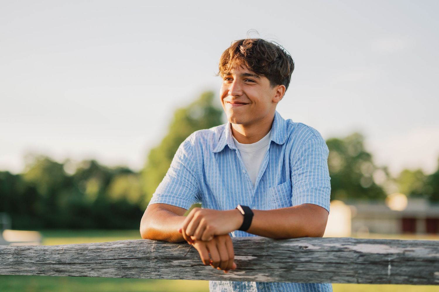 Young man leaning on wooden fence outdoors, smiling thoughtfully in golden hour sunlight wearing blue striped shirt.
