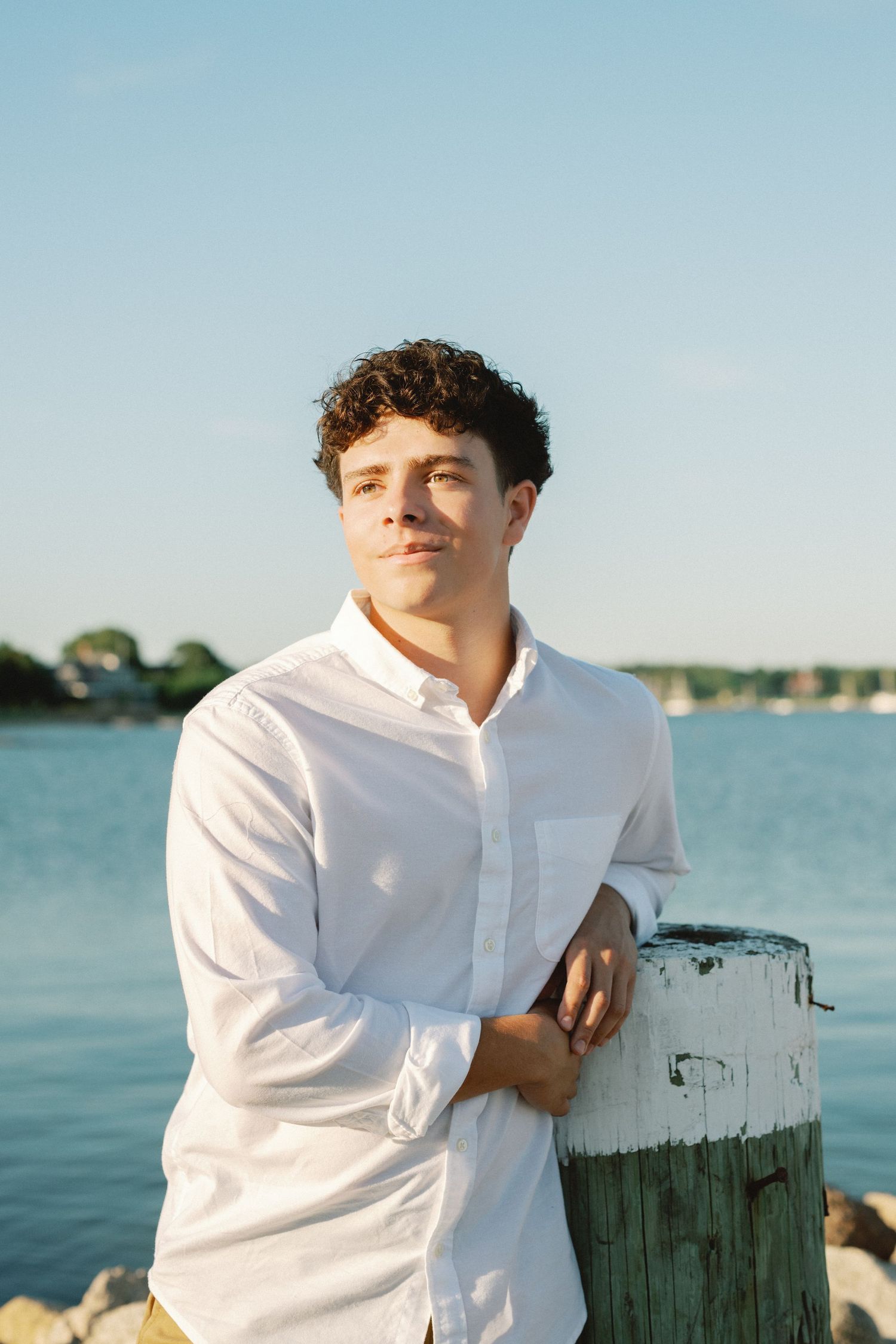 Young man in white shirt leaning on weathered dock post by the water on a sunny day.