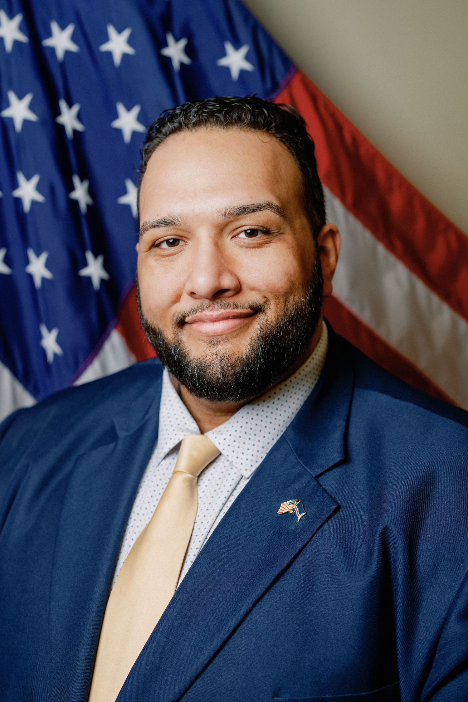 Professional headshot of a person in a navy suit and gold tie standing in front of an American flag.