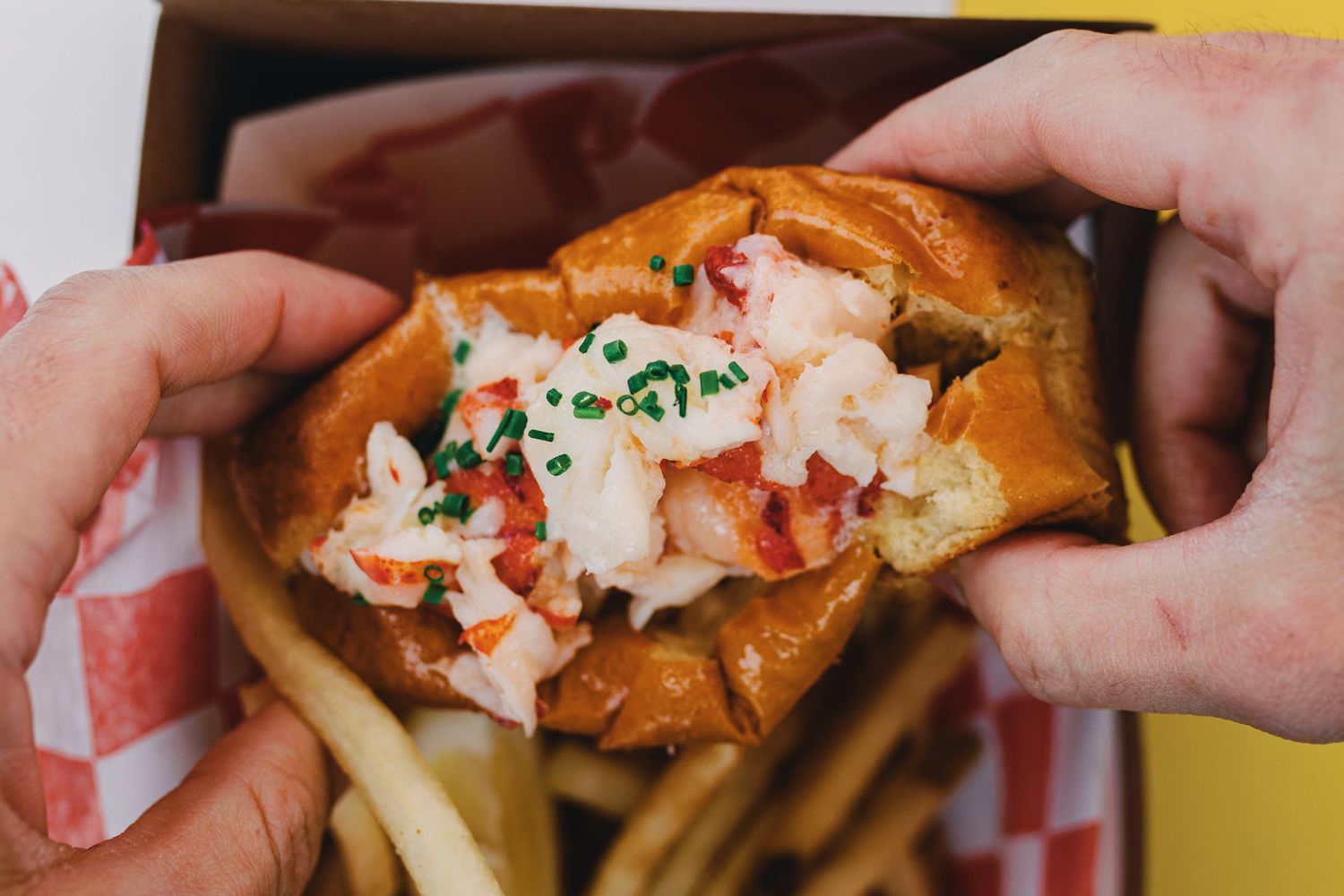 A lobster roll sandwich with chunks of meat and chives being held over french fries on a yellow background.