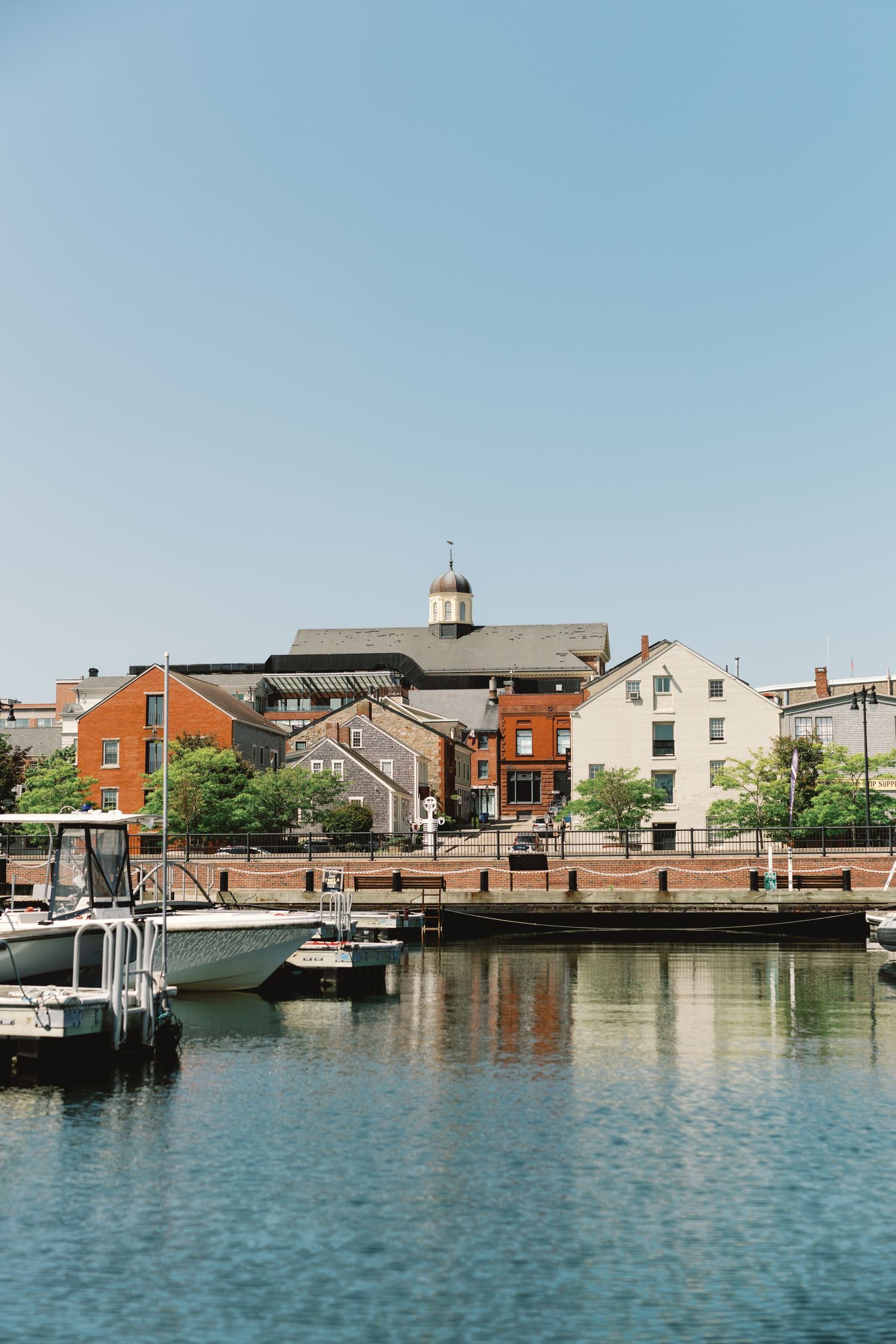 Waterfront view of historic buildings and boats along a canal with calm blue waters reflecting the sunny sky.
