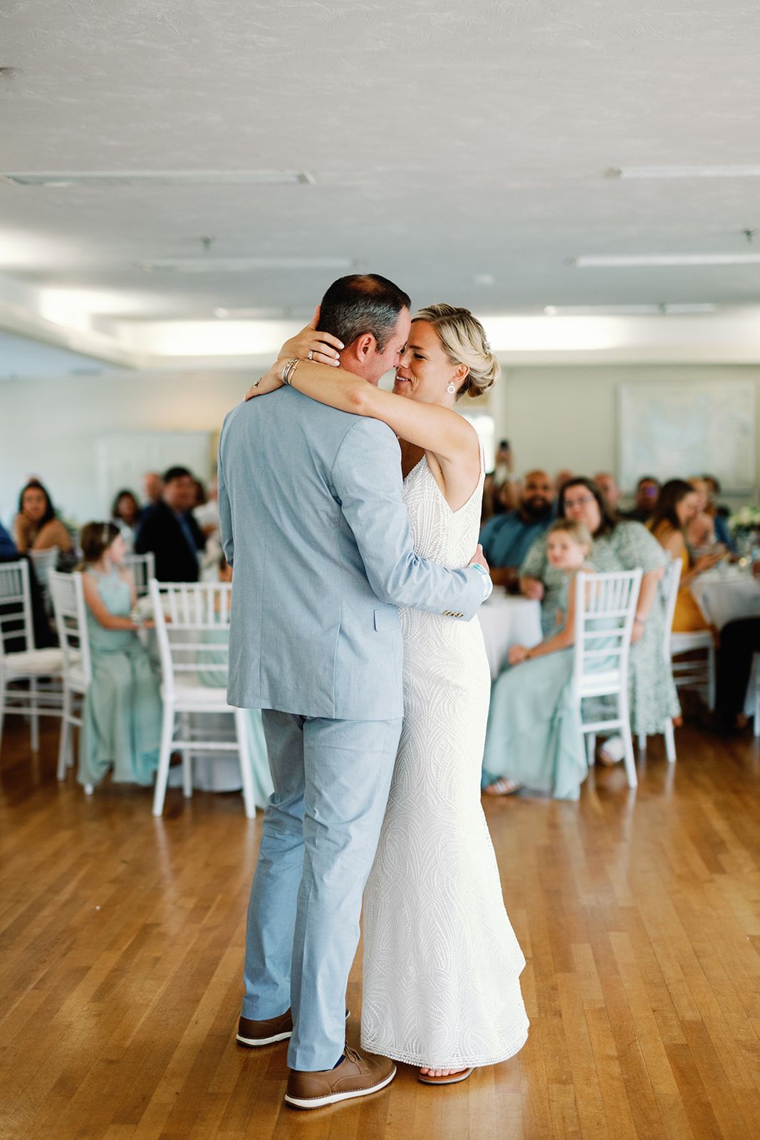 A couple shares their first dance at a wedding reception in a bright room with white chairs and wooden floors.