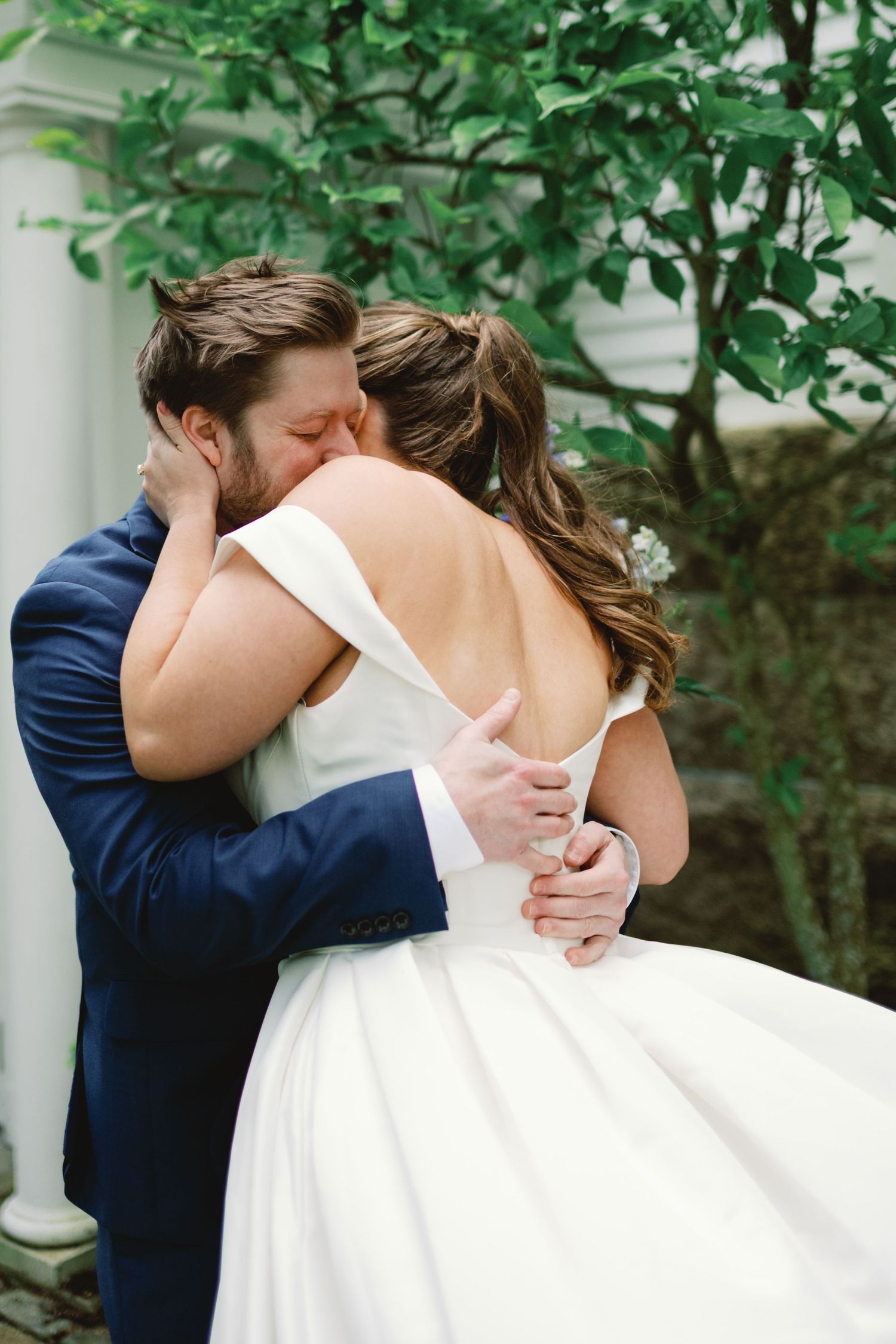 A romantic wedding moment shows a couple embracing while sharing their first kiss outdoors surrounded by greenery.