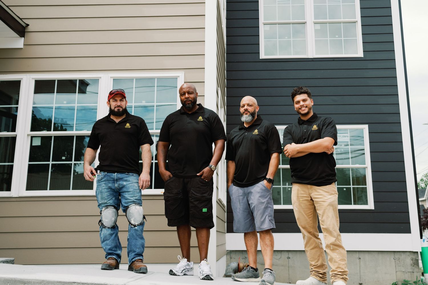 A group of construction or maintenance workers in black shirts and work pants stand in a line outside a house.