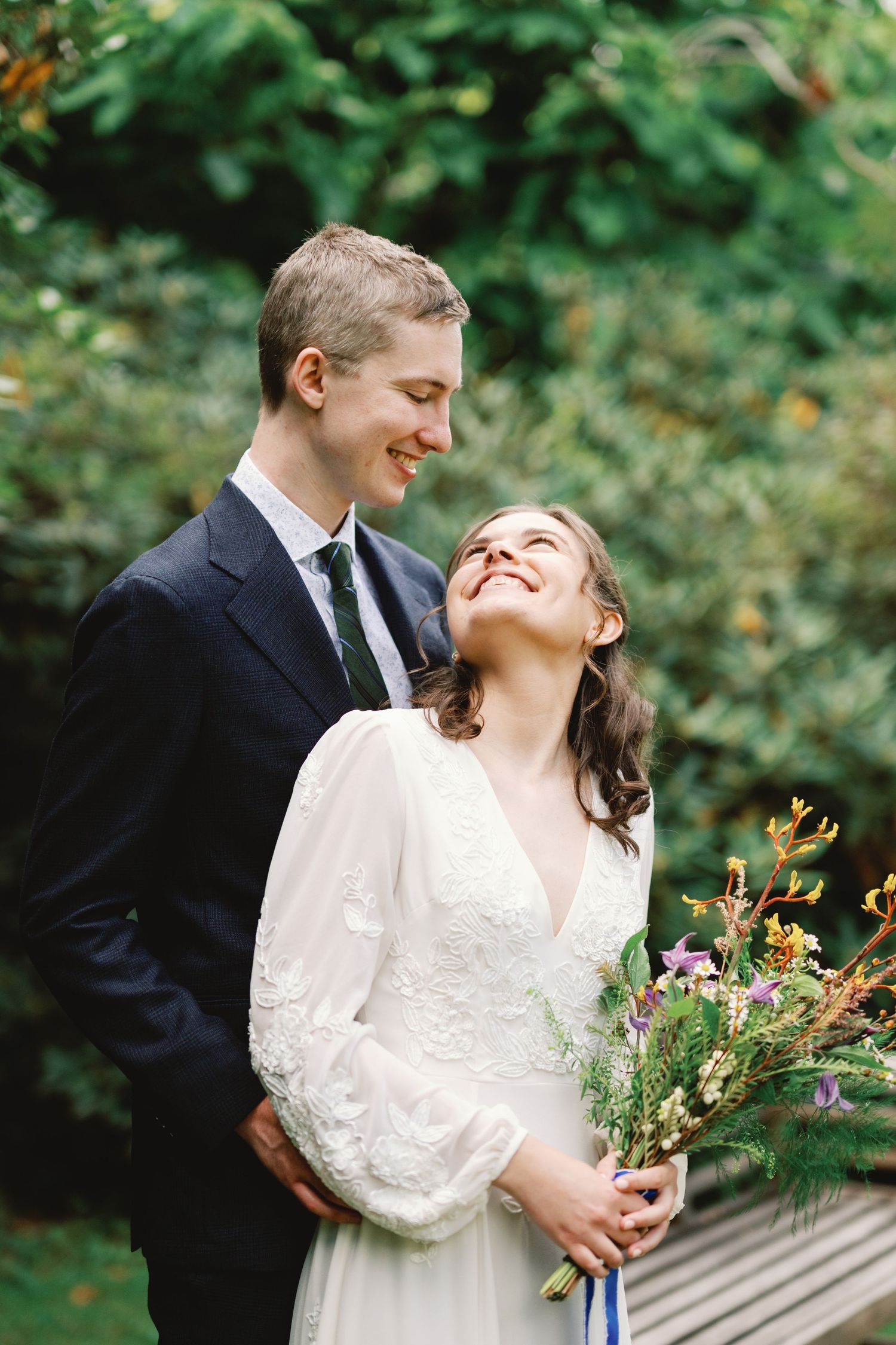 Newlyweds share a joyful moment outdoors with bride holding a wildflower bouquet in front of green foliage.