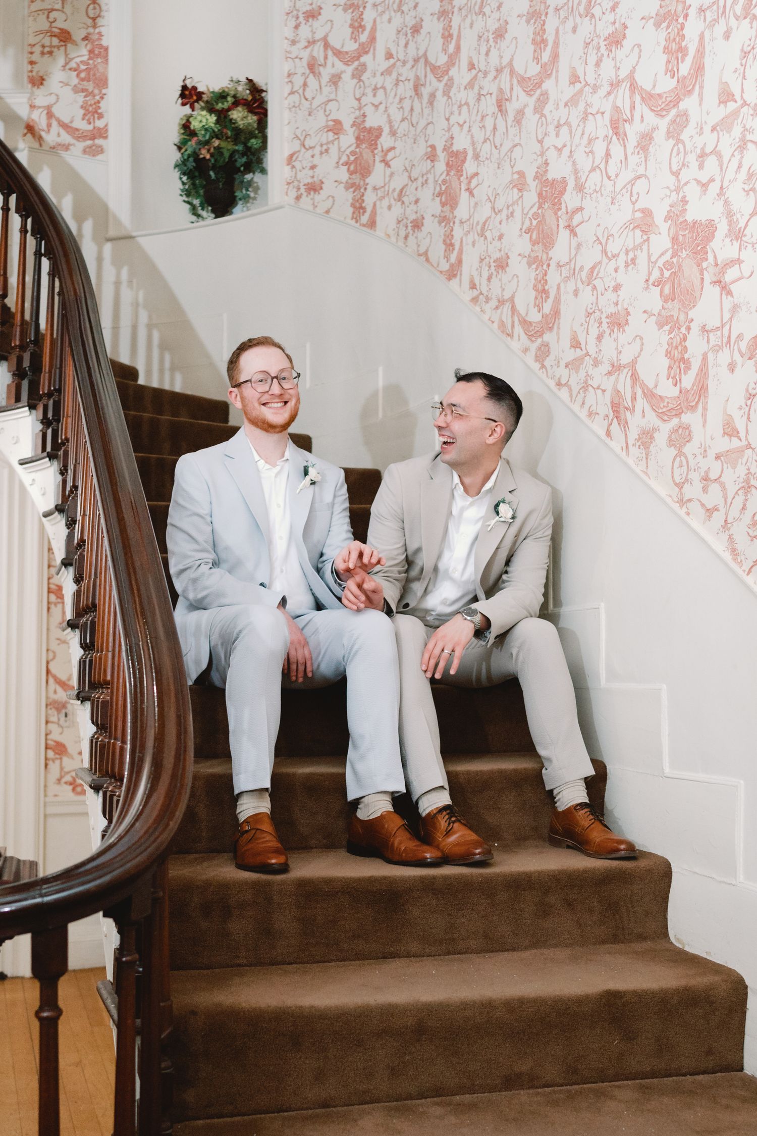 Two people in coordinating suits sitting on carpeted stairs with pink floral wallpaper behind them.