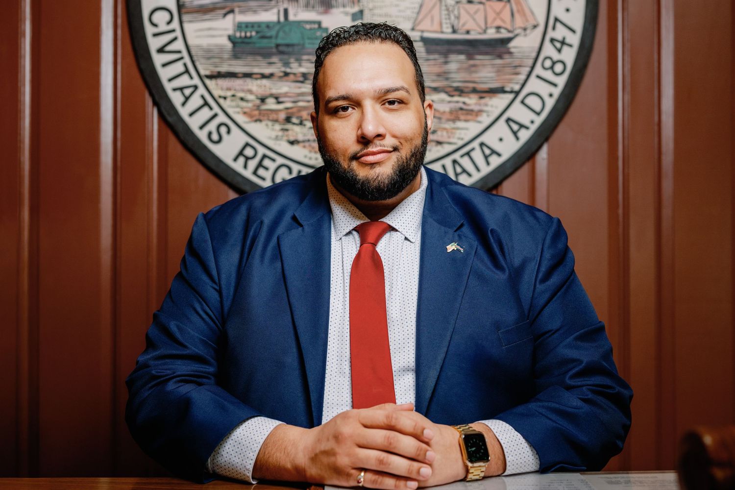 Professional man in blue suit and red tie seated at desk before official government seal backdrop.