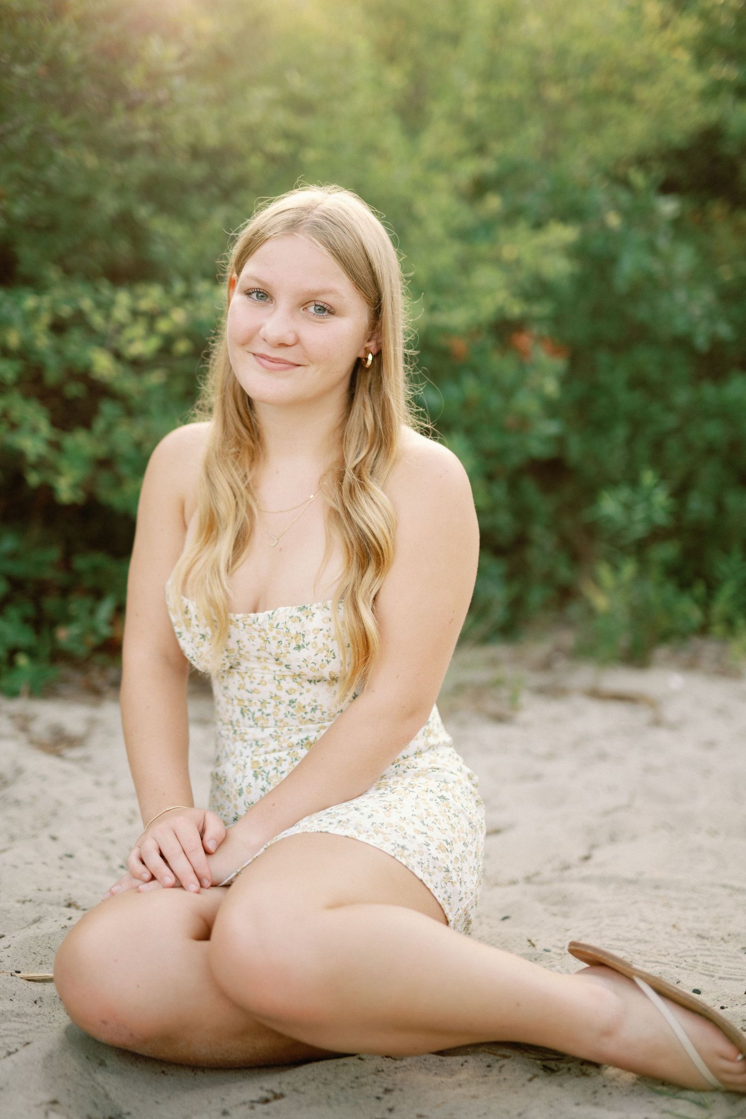 Young blonde woman in floral sundress sitting on sandy beach with green foliage background in natural light.