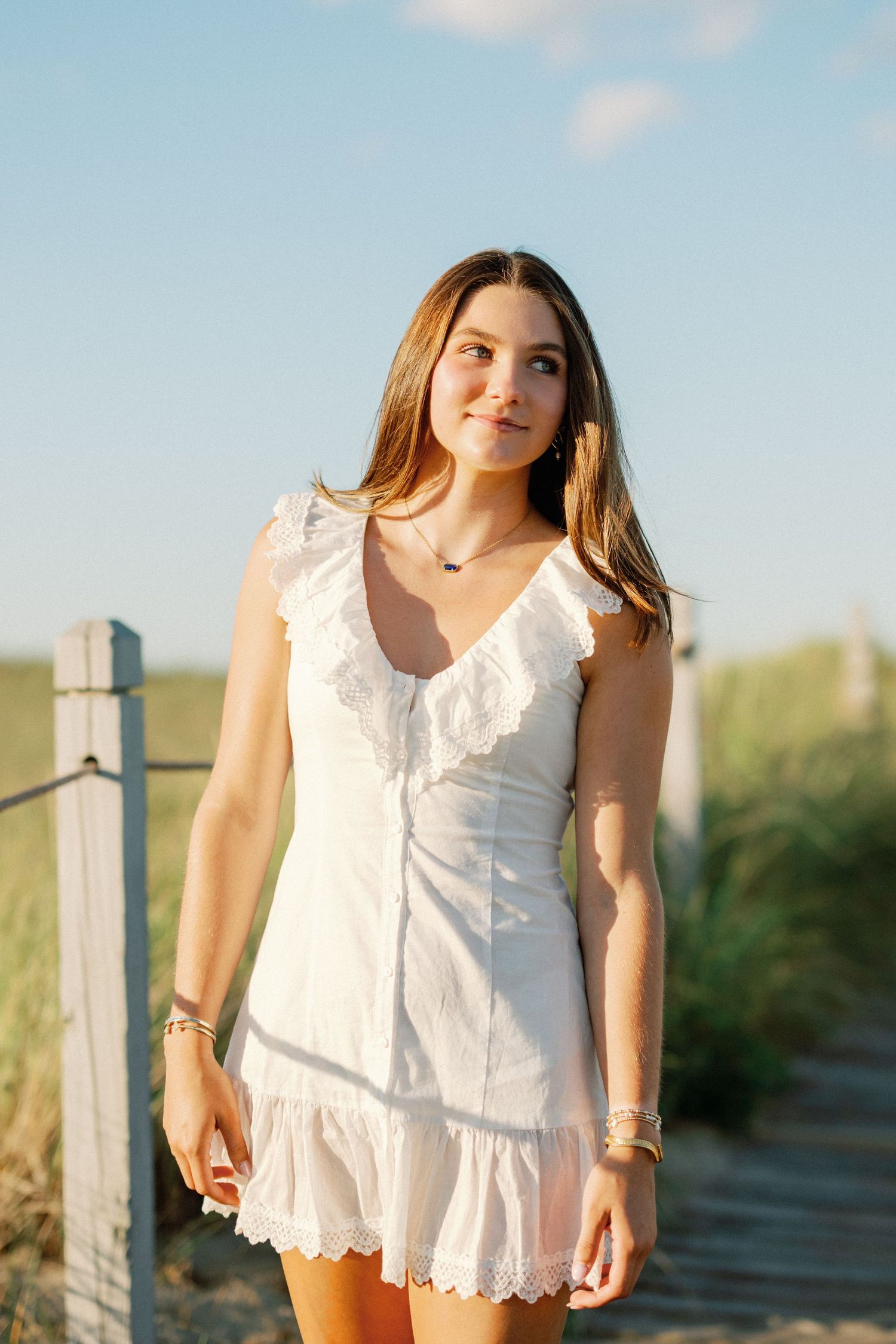 Young woman in white ruffled mini dress standing on a sunny beach path, smiling softly in golden hour light.
