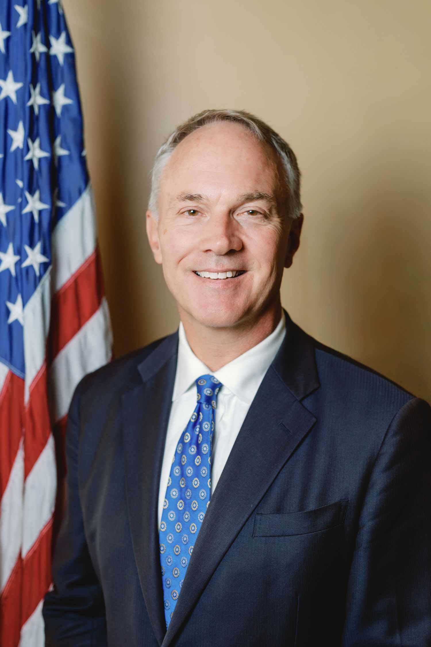 Professional headshot portrait of someone in a navy suit and blue patterned tie standing in front of an American flag.