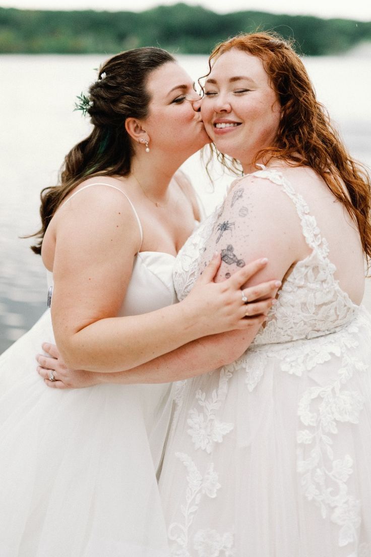 Two people in white wedding attire share an intimate embrace by a waterfront setting with trees in the background.