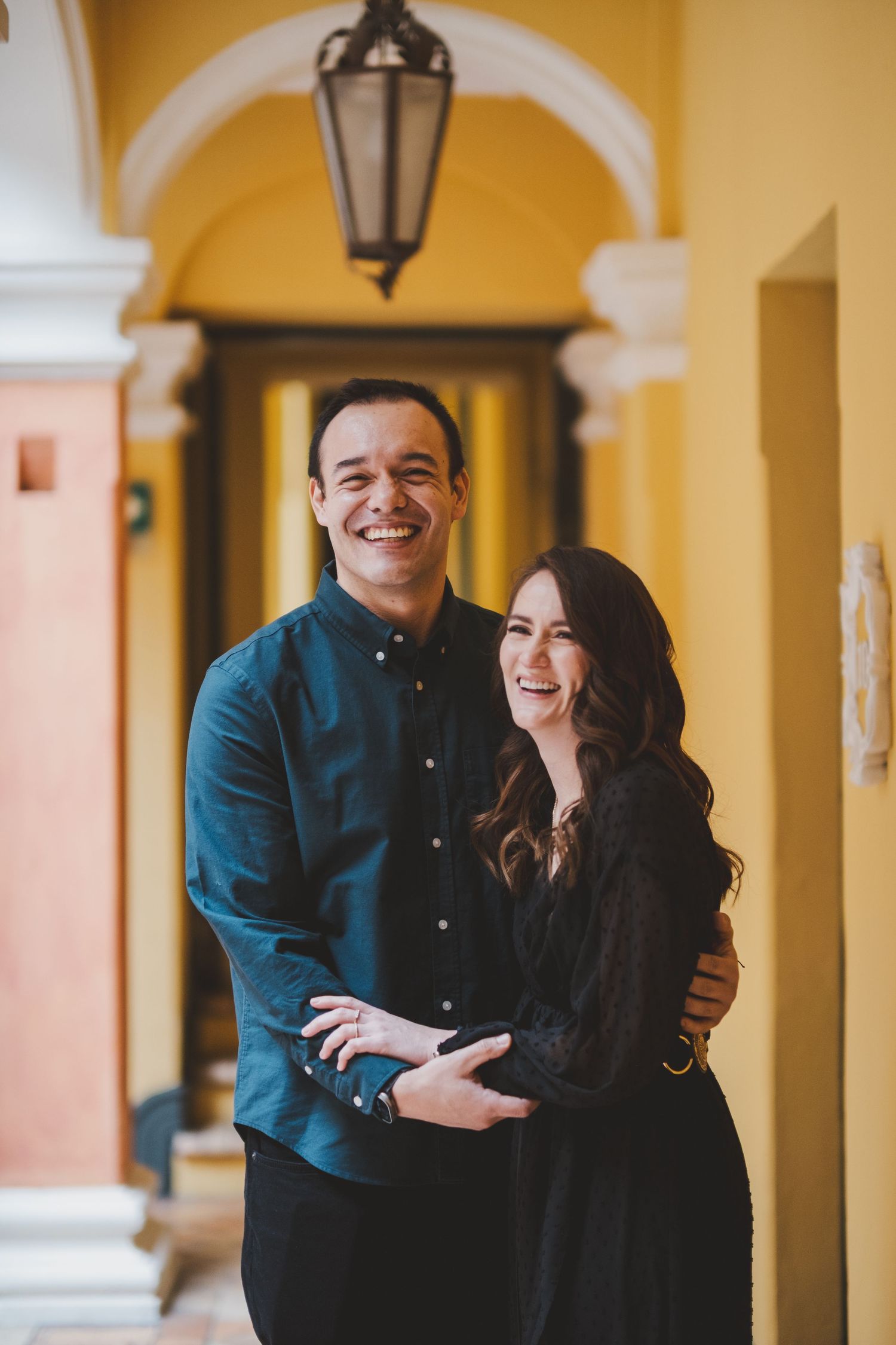A couple embraces and smiles while standing in a yellow archway corridor with decorative lantern lighting.