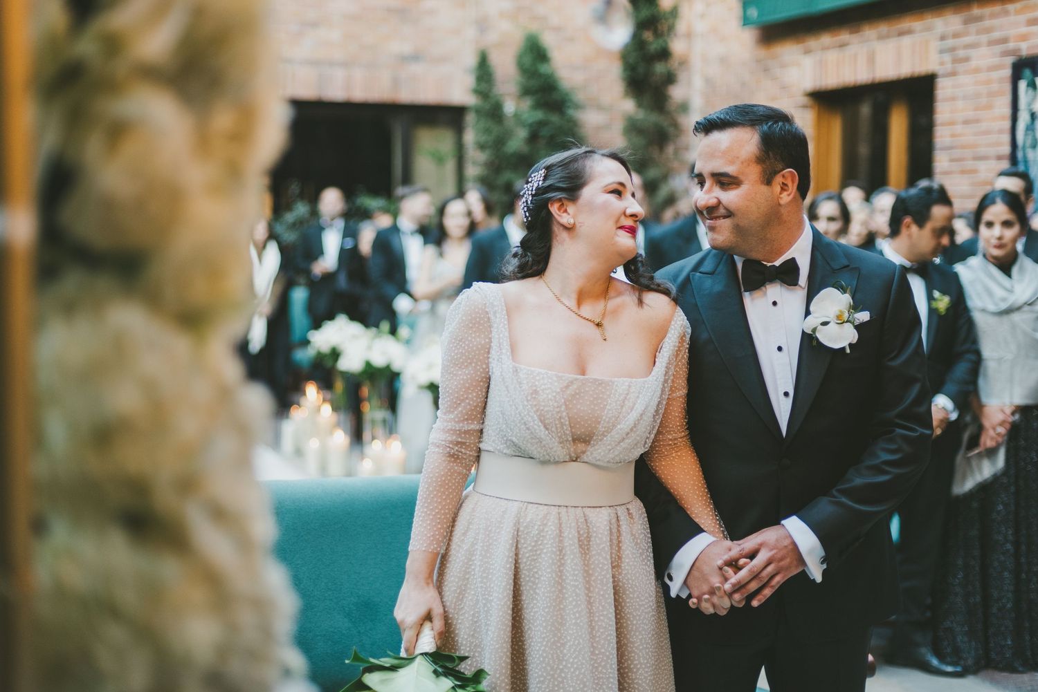 A couple joyfully walks hand in hand wearing formal attire while guests look on at an outdoor wedding venue.