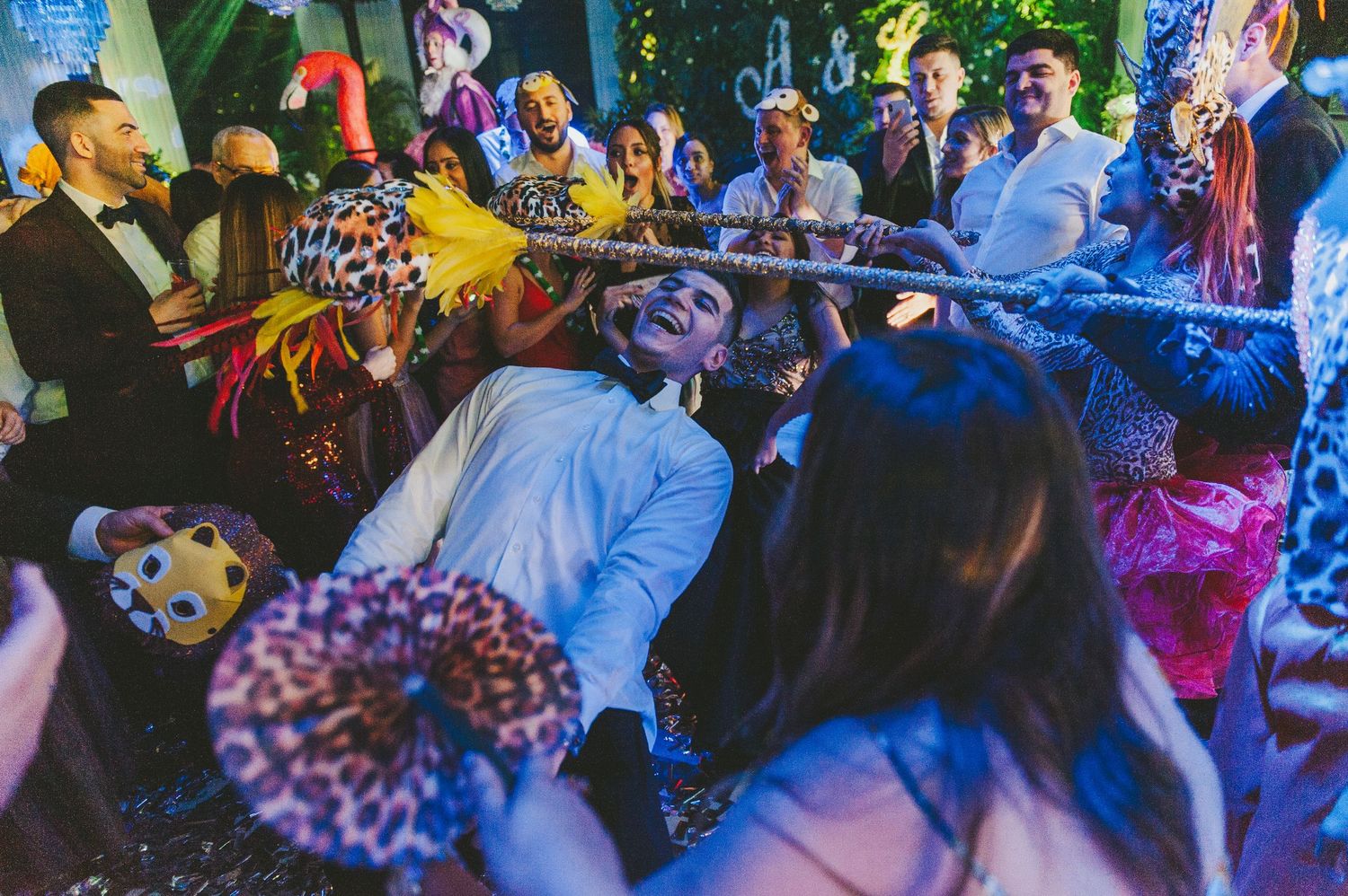 Wedding reception guests dance and celebrate beneath colorful lighting at an indoor venue during a lively party.