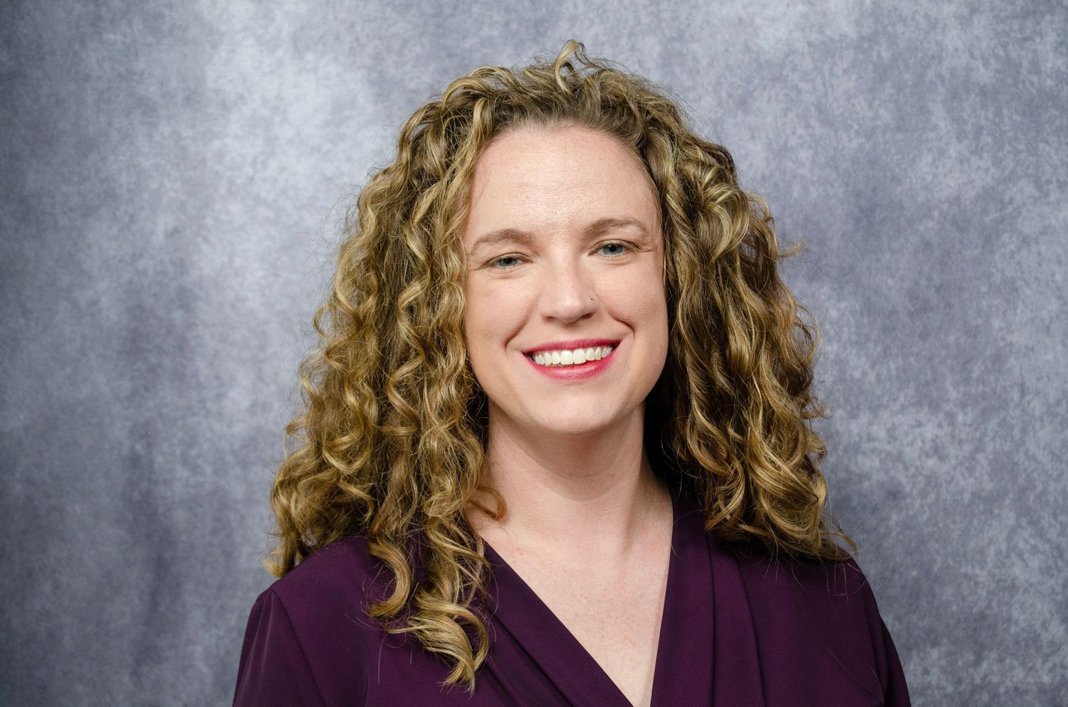 Professional headshot of someone with curly blonde hair wearing a purple top against a gray background, smiling warmly.