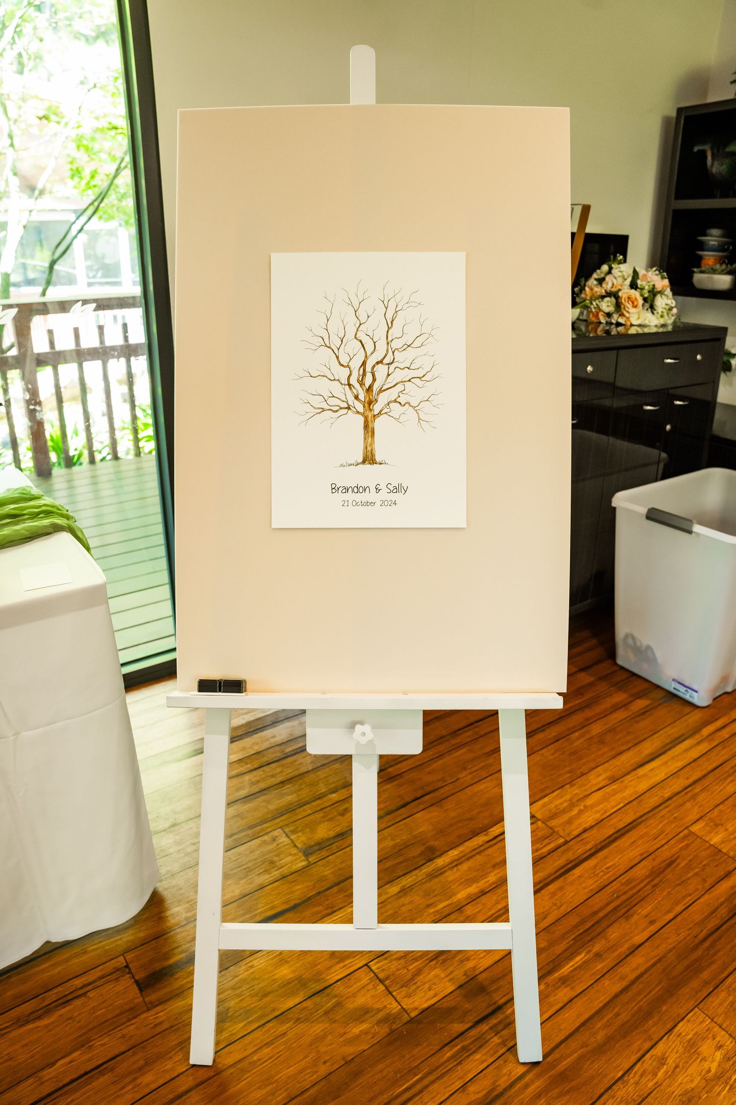 Wedding guestbook canvas displaying a bare tree artwork design sits on white easel stands on hardwood floor.