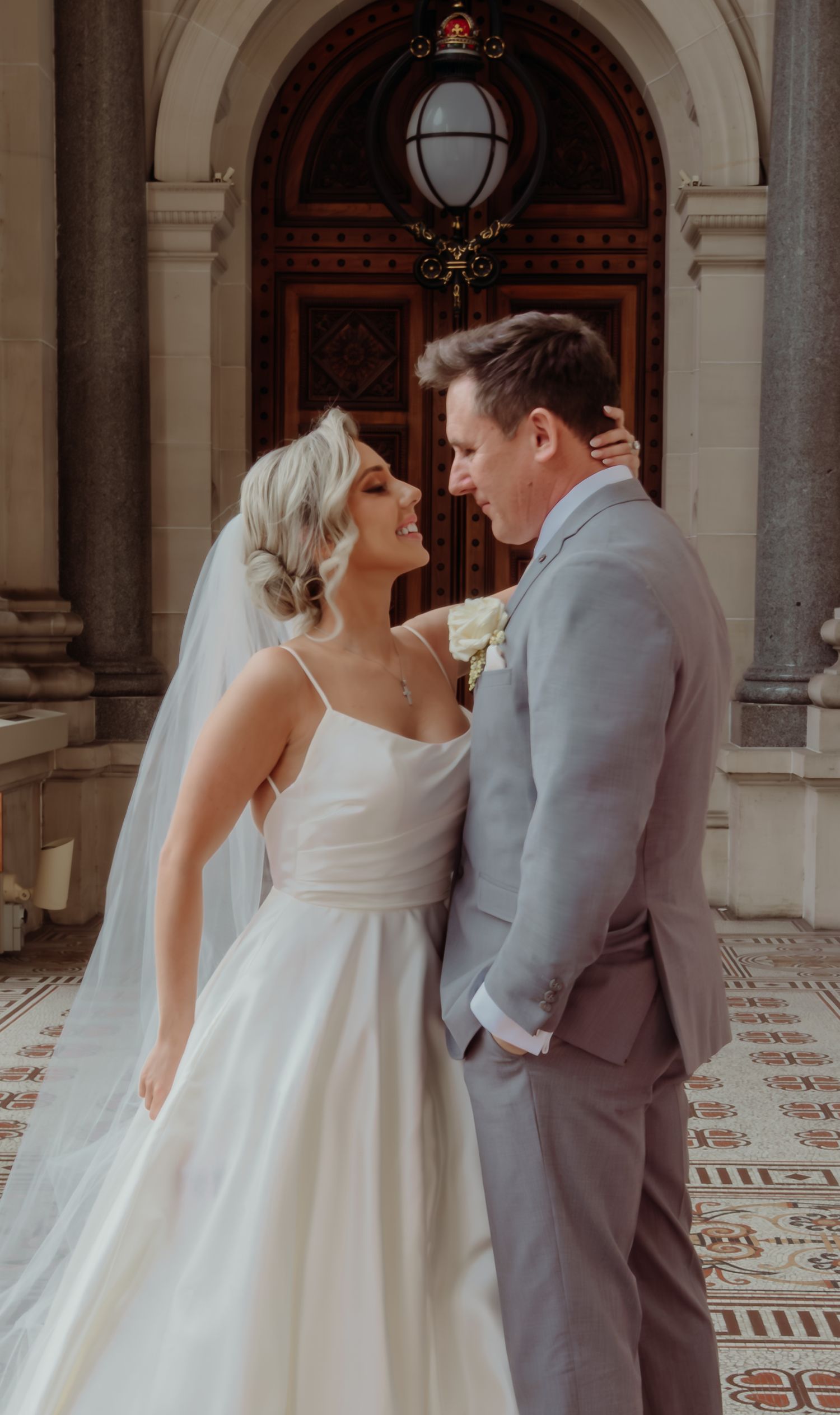A romantic wedding moment between a couple in a white gown and gray suit standing in front of an ornate doorway.