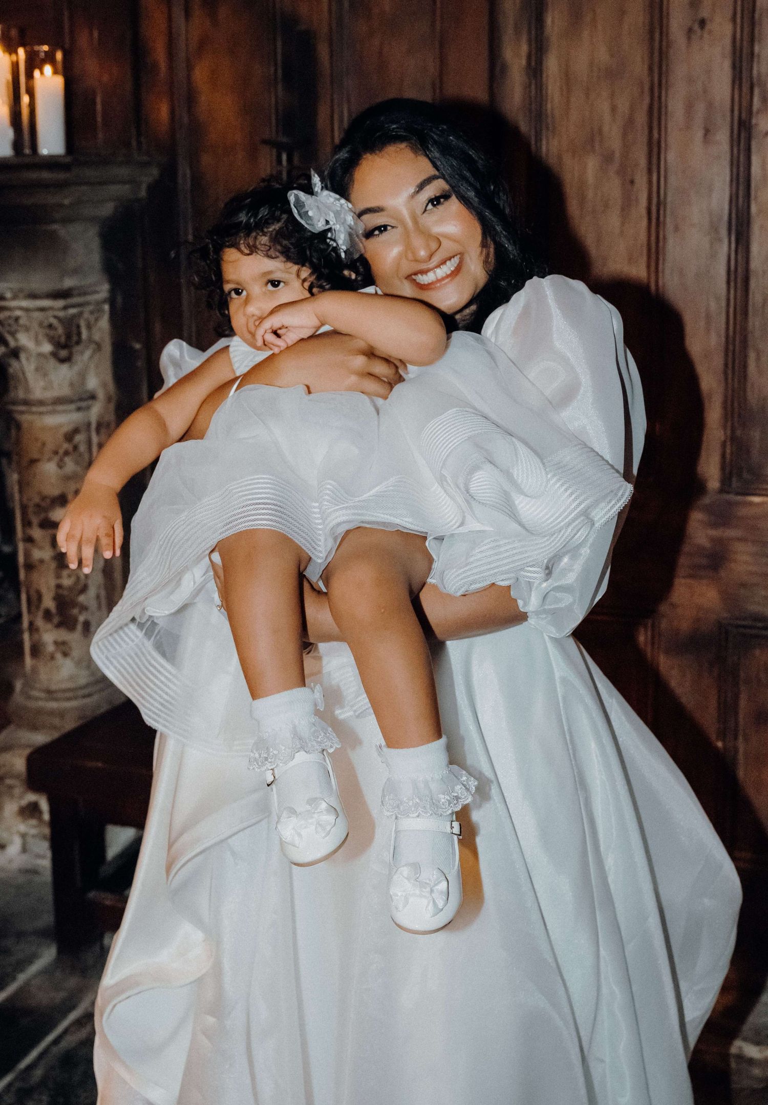 A candid moment in all white attire showing a tender embrace between adult and child against a rustic wooden backdrop.
