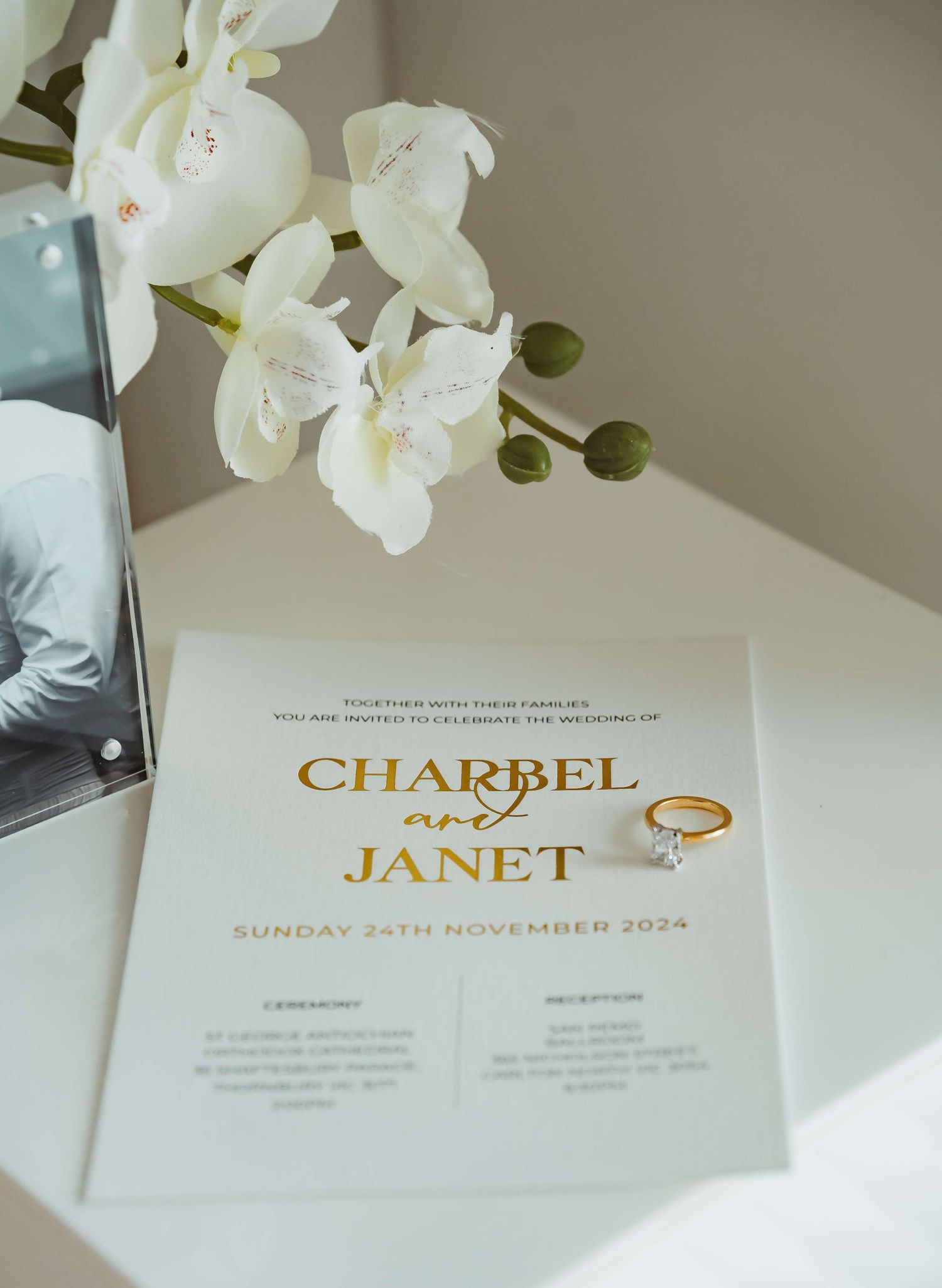 An elegant wedding invitation with gold text for Charbel and Janet sits beside white orchids and buds in soft lighting.
