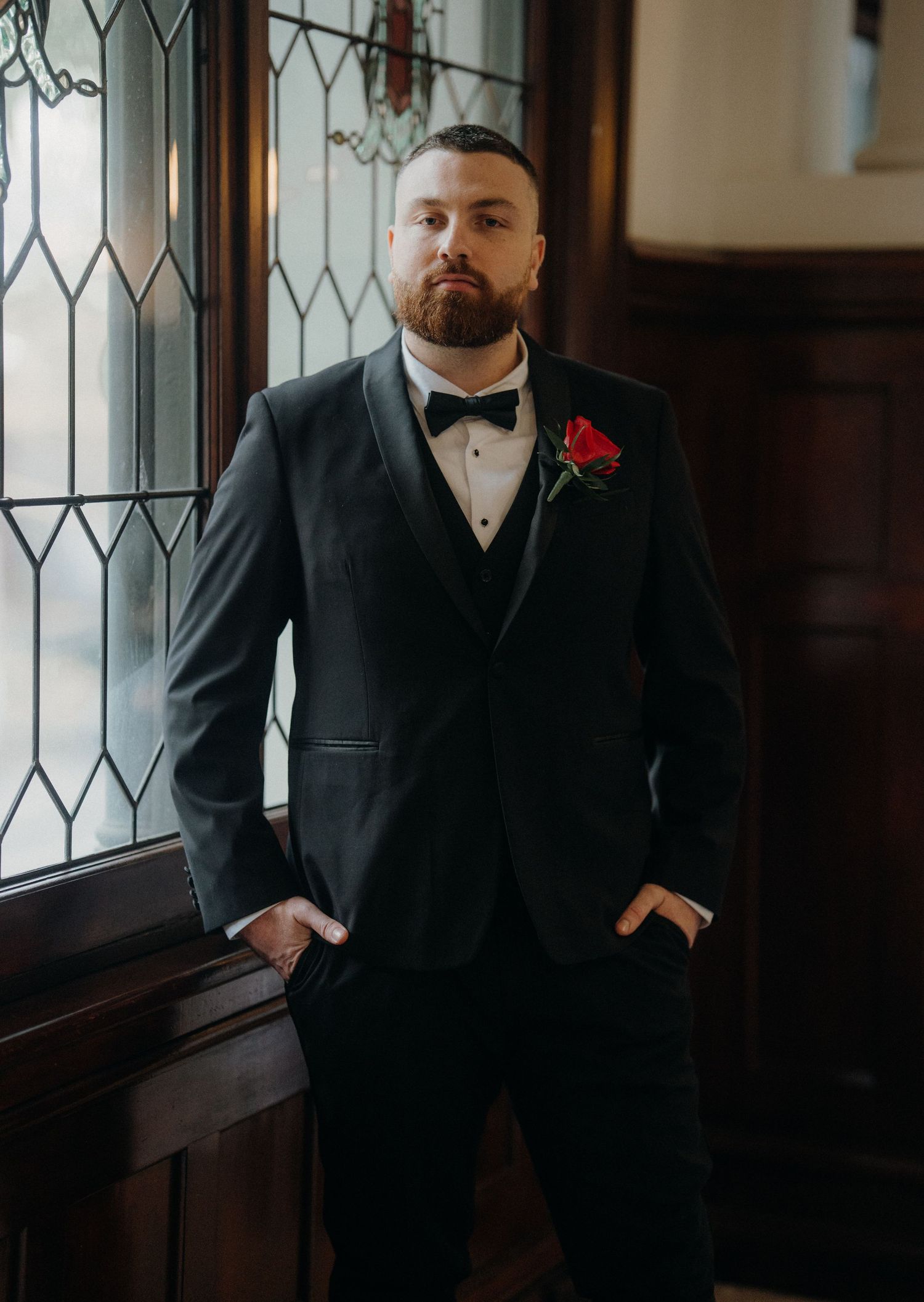 Groom in black tuxedo with bow tie and red rose boutonniere stands by stained glass window.