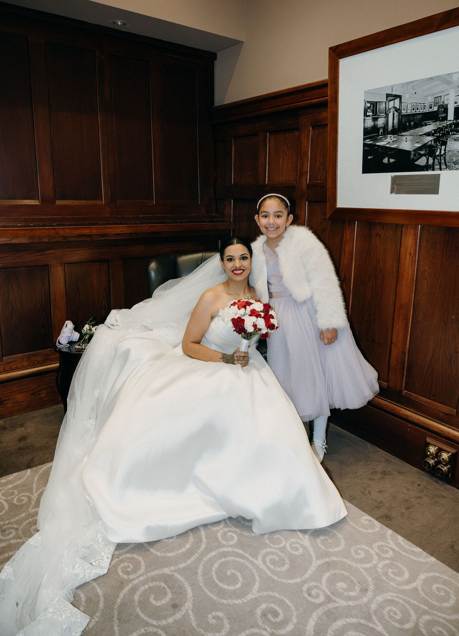 Bride in white gown holding red bouquet poses with young flower girl in lavender dress and white fur shrug indoors.