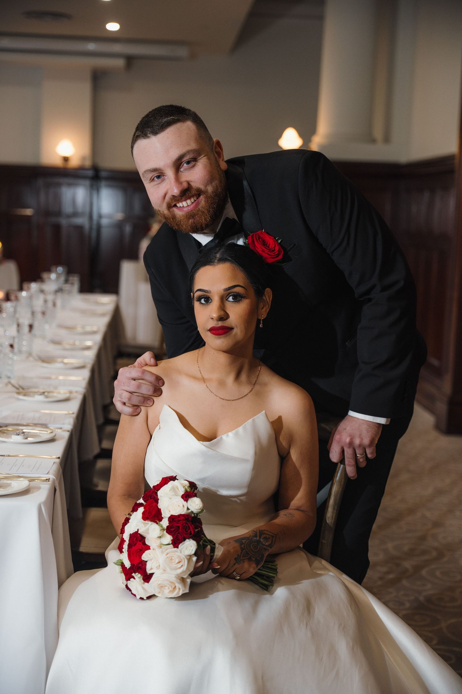 Bride in white gown holding red and white bouquet sits as groom in black tuxedo with red rose stands behind her.